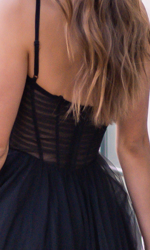 A person wearing a black dress is seen from the back. The dress features a fitted bodice with vertical stripes, leading into a flowing skirt, complemented by wavy hair cascading down.