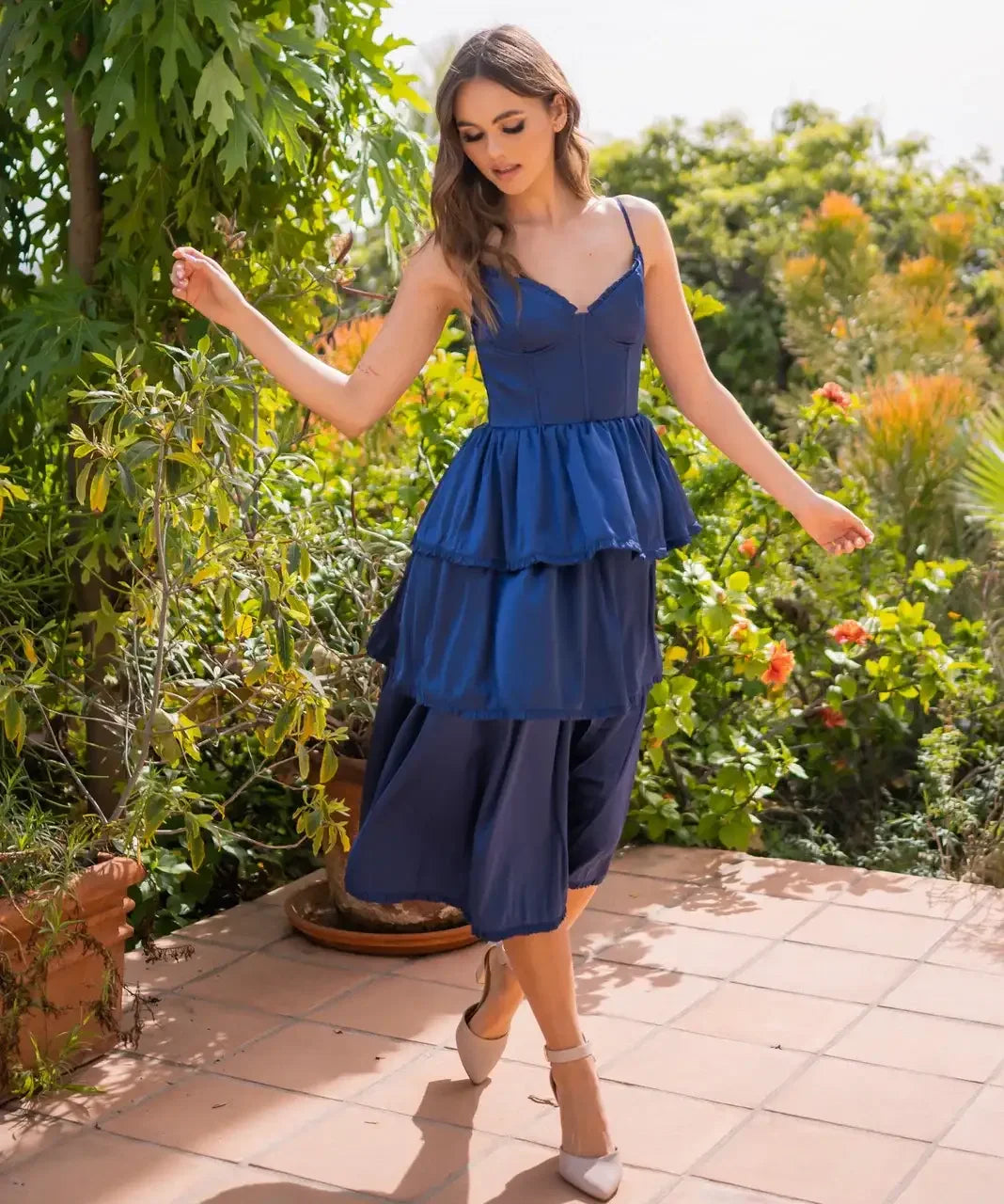 A woman in a blue dress with layered ruffles gracefully walks on a tiled patio, surrounded by lush greenery and colorful flowers under a bright, sunny sky.