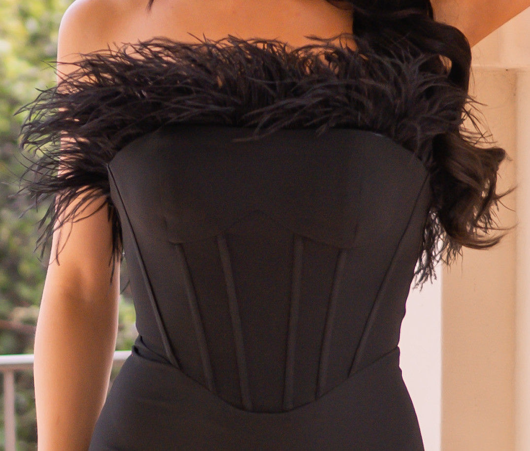 A black strapless outfit features a feathered neckline, creating a dramatic appearance. The wearer stands against a light-colored wall, with greenery visible in the background.