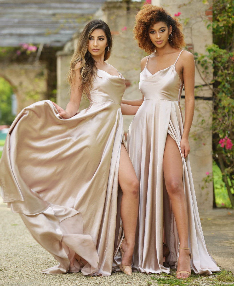 Two women stand side by side, wearing elegant, flowing satin dresses with high slits. They pose confidently in a lush garden setting, surrounded by greenery and blooming flowers.