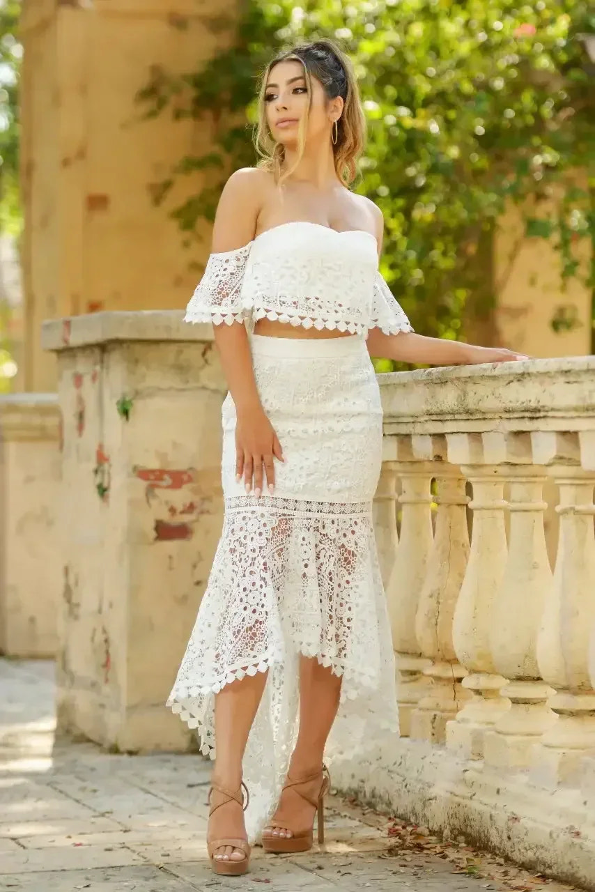A woman stands gracefully beside a stone railing, wearing a white lace two-piece outfit. The setting features lush greenery and weathered architecture on a sunny day.
