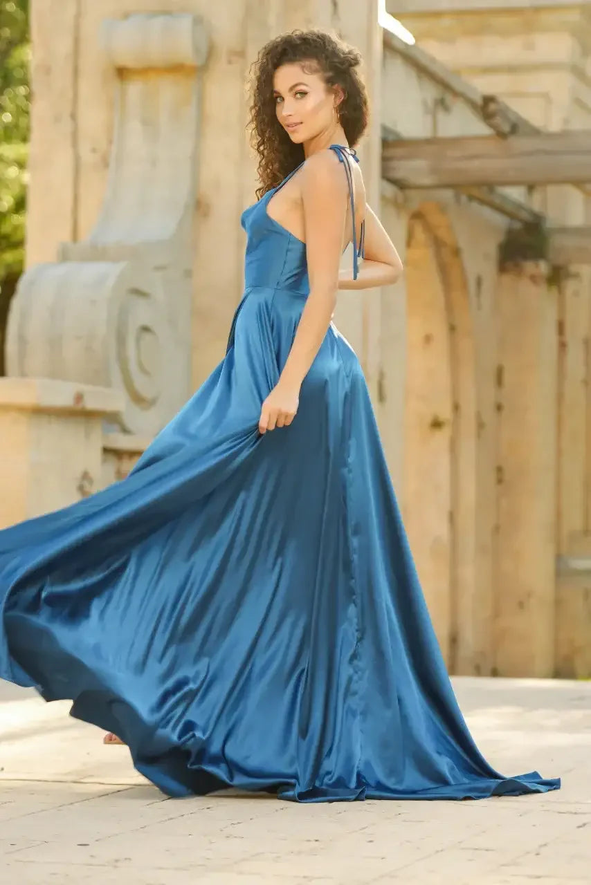 A woman in a flowing blue satin dress gracefully turns, showcasing the gown's movement. The scene is set in an outdoor location with stone architecture and natural lighting.