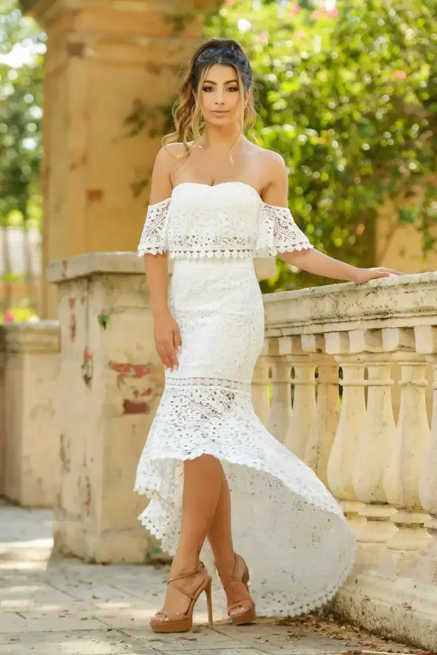 A woman in a white, lace-off-shoulder dress stands elegantly near a stone railing in a garden. Sunlight filters through lush greenery, enhancing the serene, natural atmosphere.