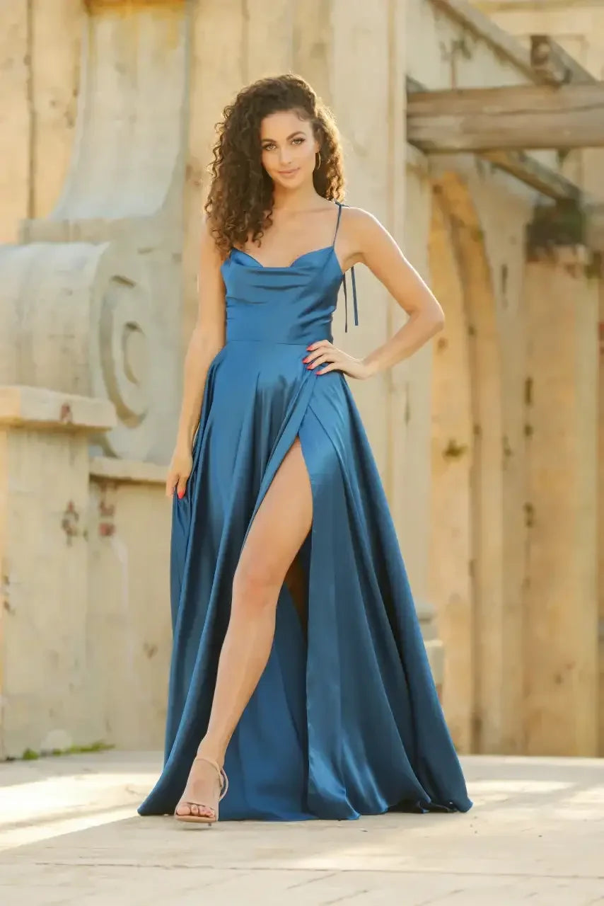 A woman in a flowing, blue satin gown with a high slit stands confidently on a sunlit stone surface. The backdrop features textured walls and wooden beams, suggesting an elegant outdoor setting.
