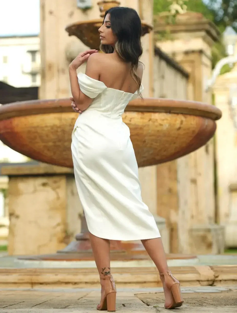 A woman in a white off-the-shoulder dress stands gracefully by a fountain, gazing back over her shoulder, with greenery and stone structures in the background.