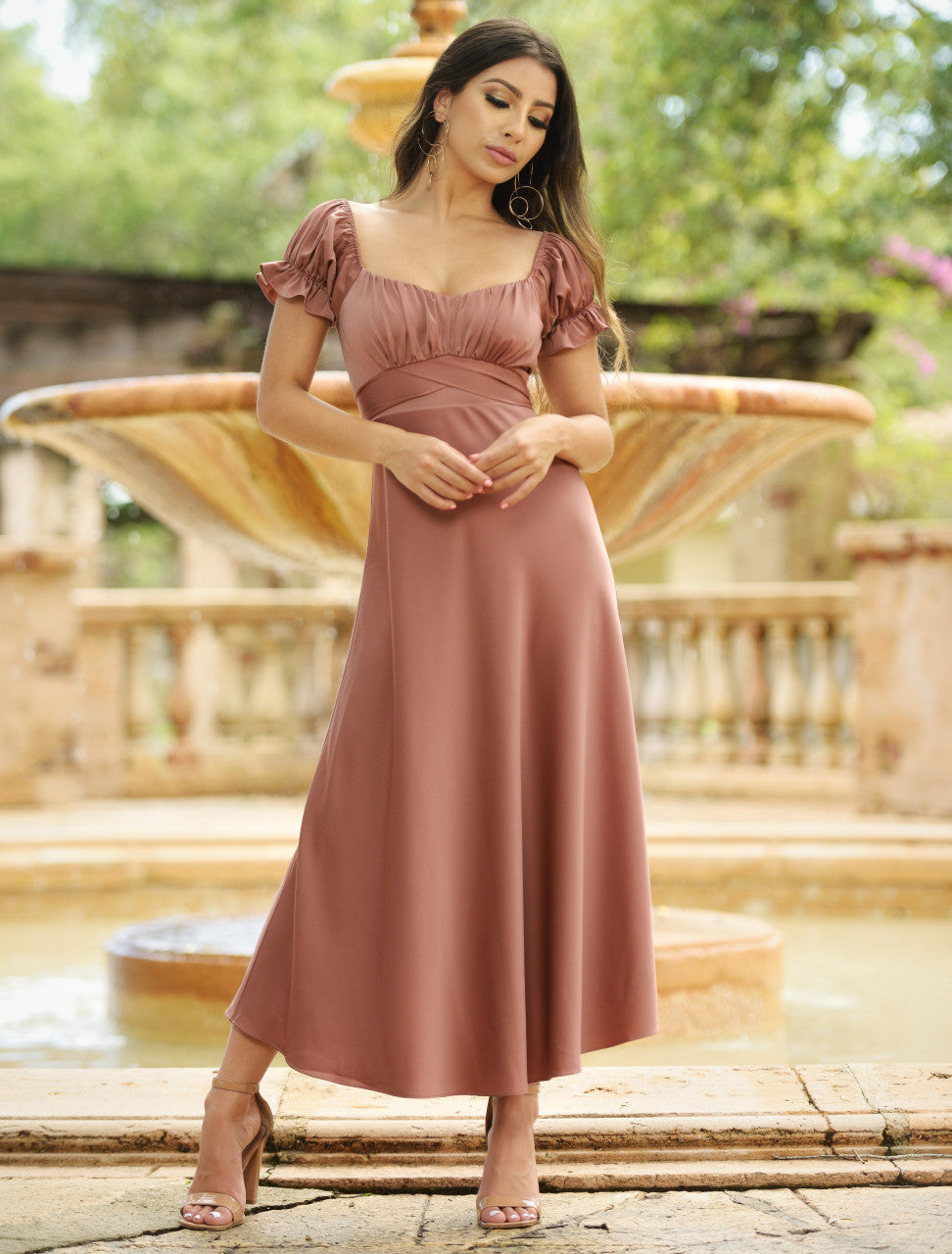 A woman in a long, mauve dress stands gracefully beside a fountain in a lush garden, her hair flowing and posture poised, creating an elegant atmosphere.