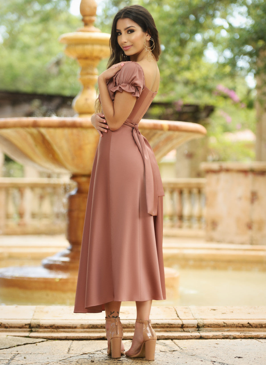 A woman in a flowing, dusty rose dress stands beside a fountain, gazing over her shoulder. The serene outdoor setting features lush greenery and elegant stone architecture in the background.