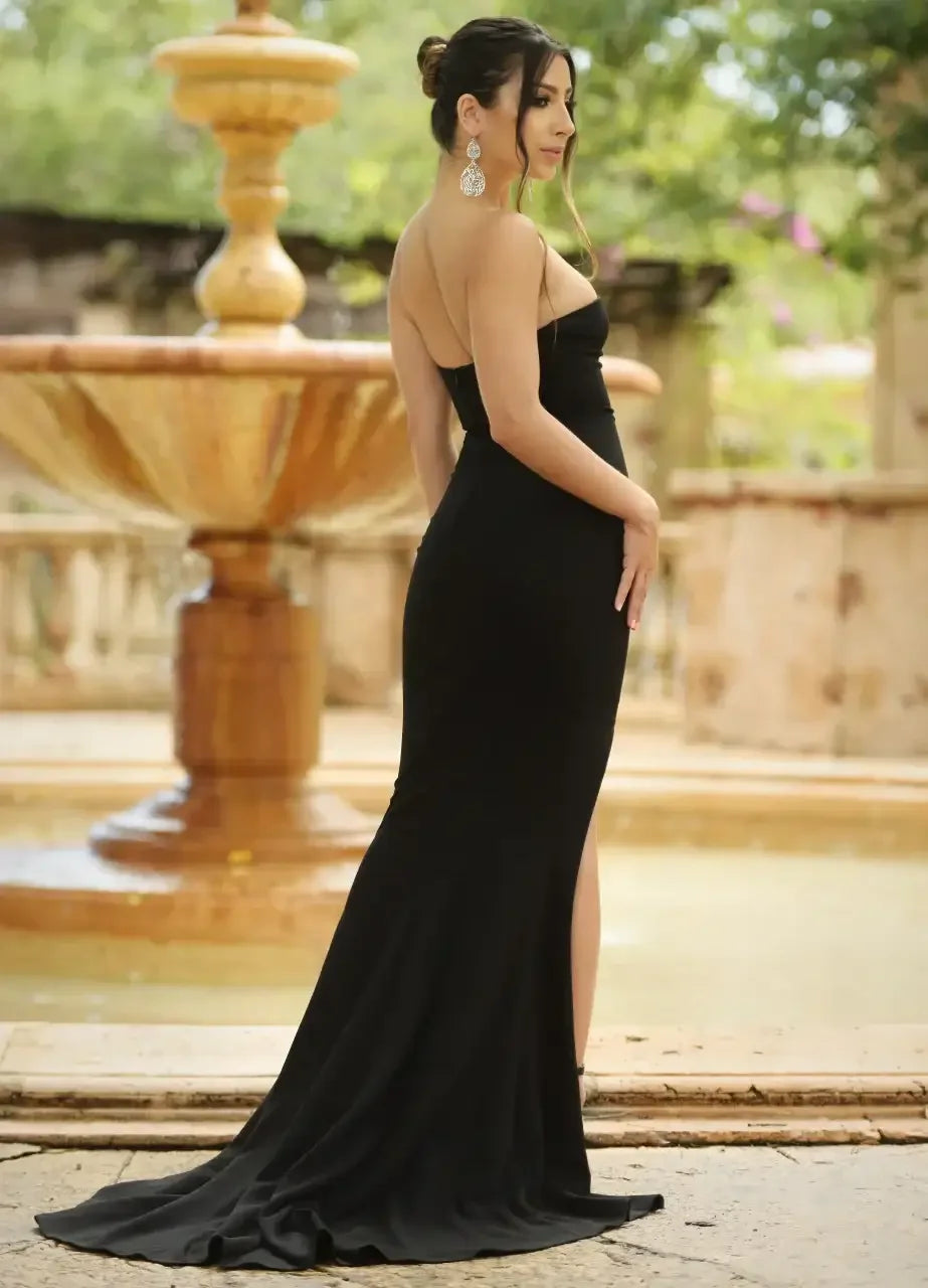 A woman in a black evening gown stands near a decorative fountain, showcasing her elegant posture and long hair, with a blurred garden setting and water feature in the background.