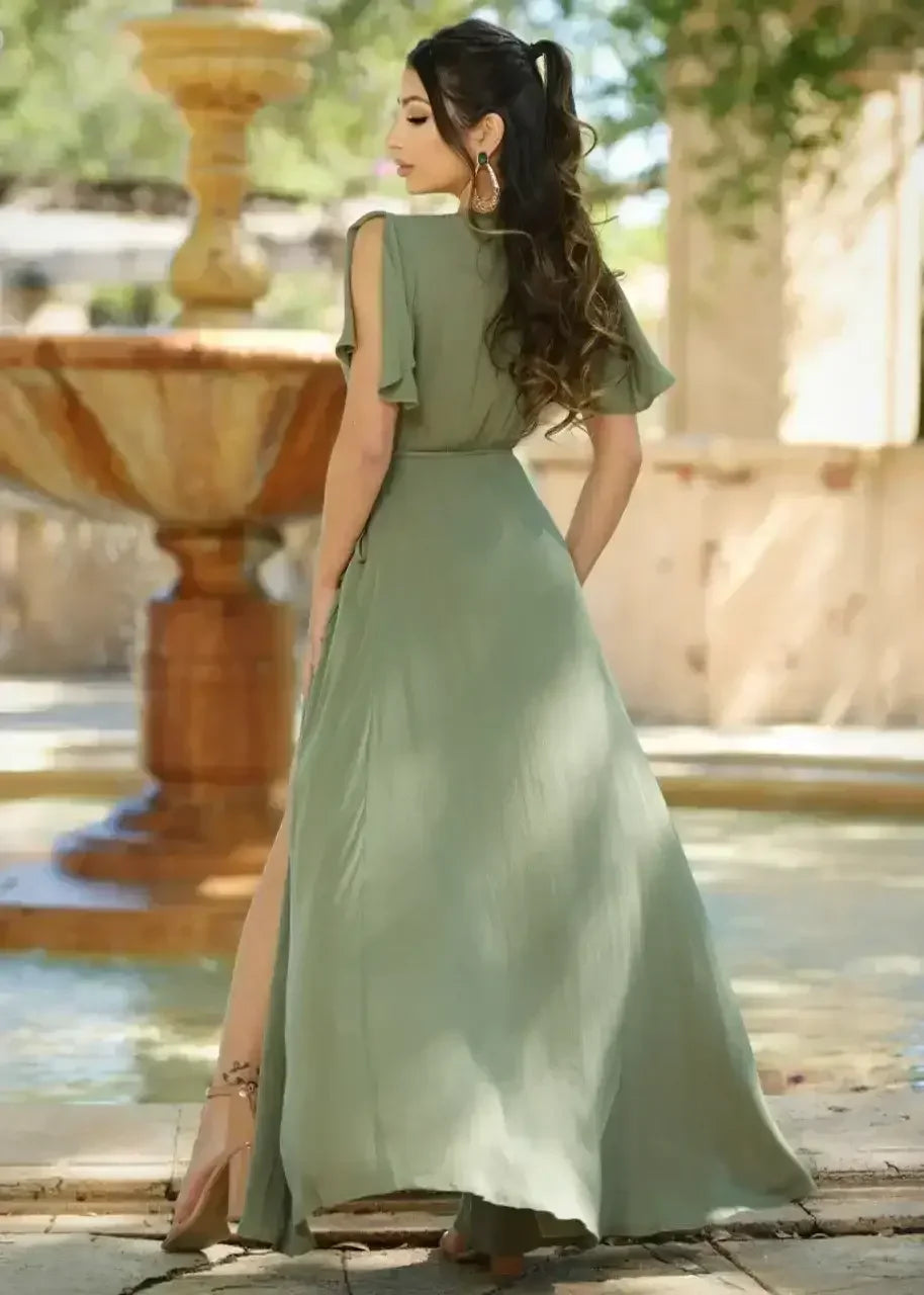A woman in a long, olive green dress stands gracefully with her back turned, facing a stone fountain in a serene outdoor setting, surrounded by greenery and soft sunlight.