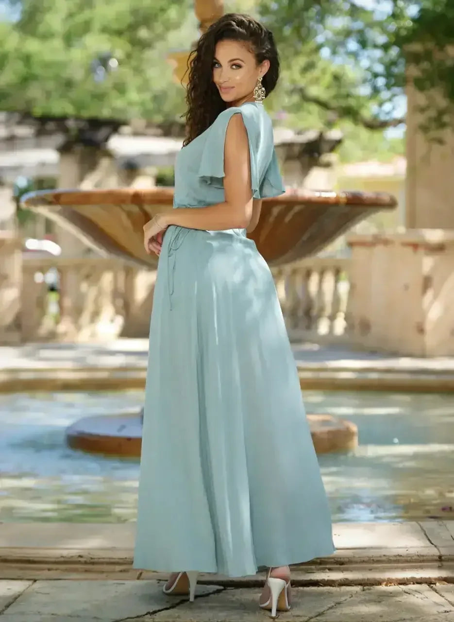A woman in a light blue, flowing dress stands by a decorative fountain, posing gracefully with a slight smile. The scene is set in a garden with lush greenery.