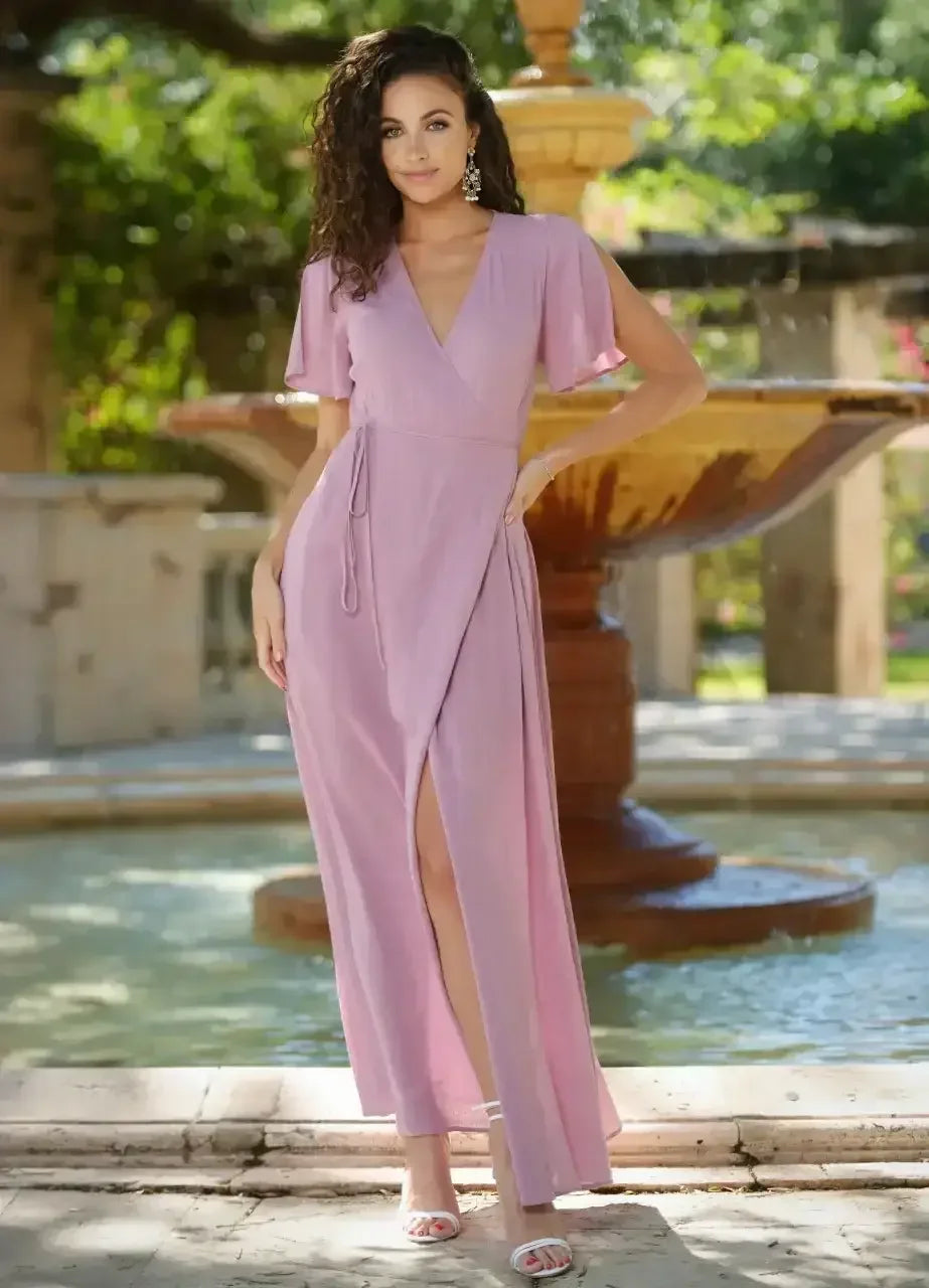 A woman wearing a flowing, pink maxi dress with short sleeves stands next to a fountain in a lush garden, smiling softly. Sunlight filters through the greenery.