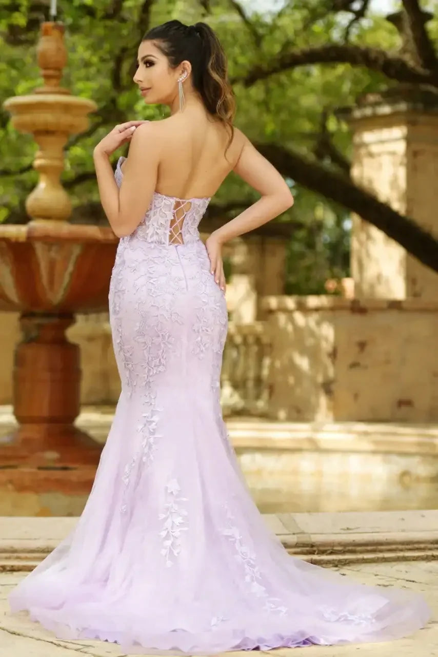 A woman in a lavender strapless gown with intricate floral embroidery poses gracefully, showcasing the gown's fitted silhouette and flowing train. She stands near a decorative fountain in a lush garden setting.
