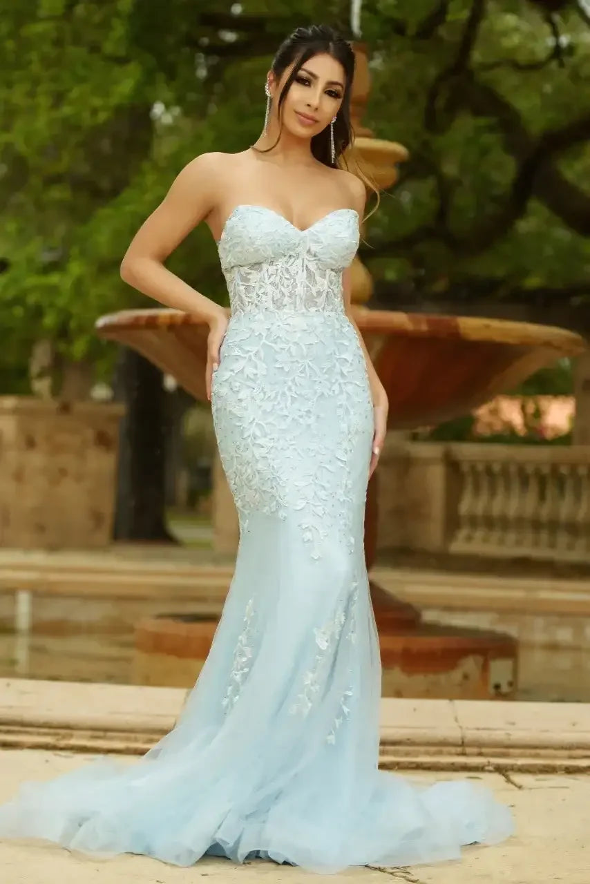 A woman in a light blue, form-fitting dress with floral lace details poses confidently near a stone fountain surrounded by greenery, captured in a vibrant outdoor setting.