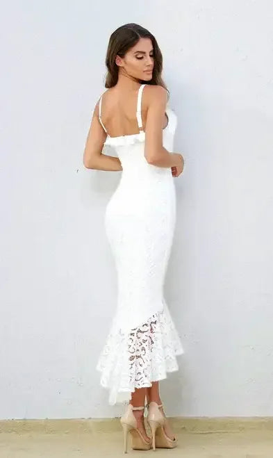 A woman stands wearing a form-fitting white lace dress with a flared hem. She is turned slightly to the side, showcasing her posture against a plain light-colored background.