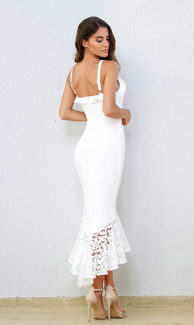 A woman stands wearing a form-fitting white lace dress with a flared hem. She is turned slightly to the side, showcasing her posture against a plain light-colored background.