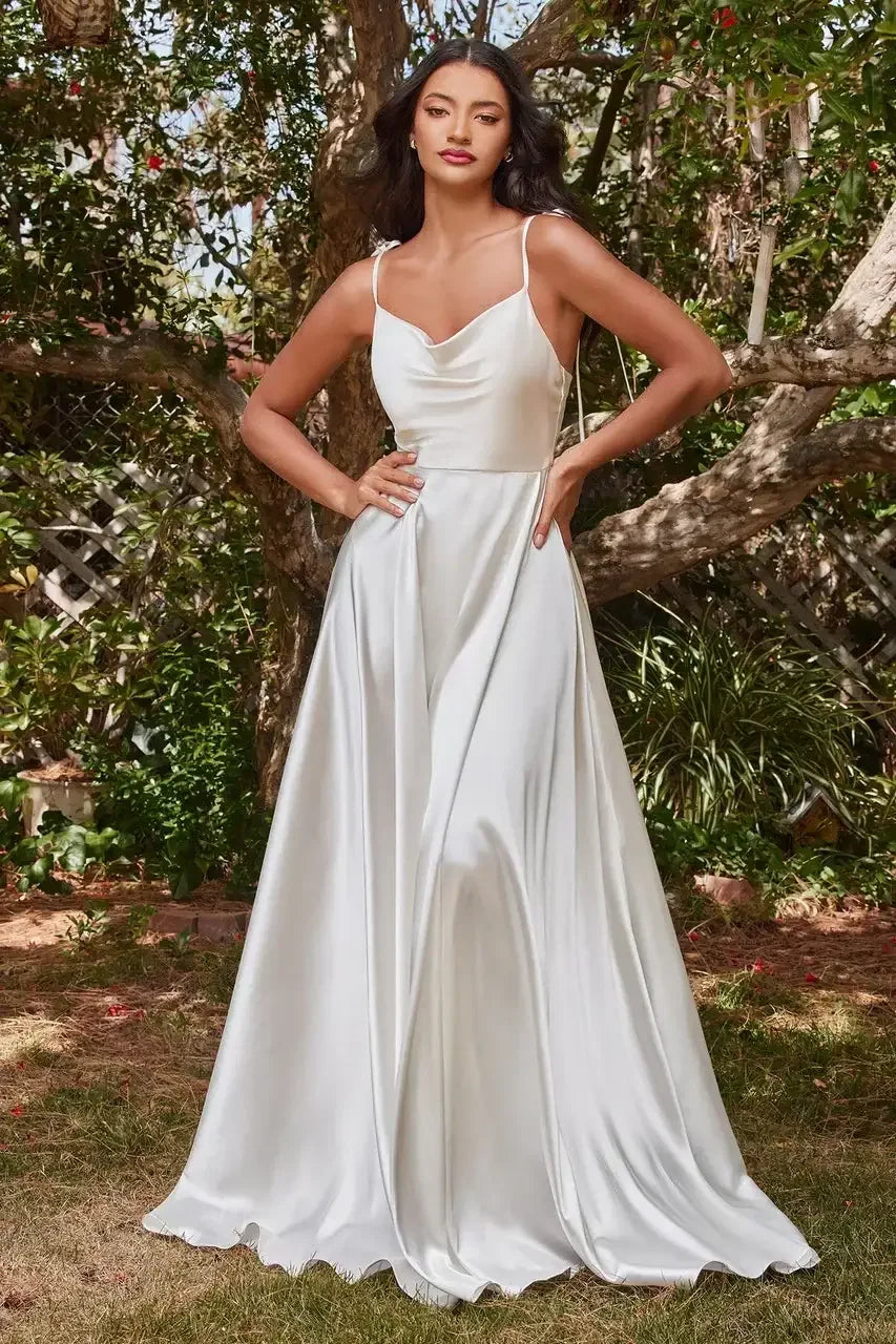 A woman poses confidently in a flowing white satin dress, showcasing its elegant drape. She stands in a lush, green outdoor setting, surrounded by trees and foliage.