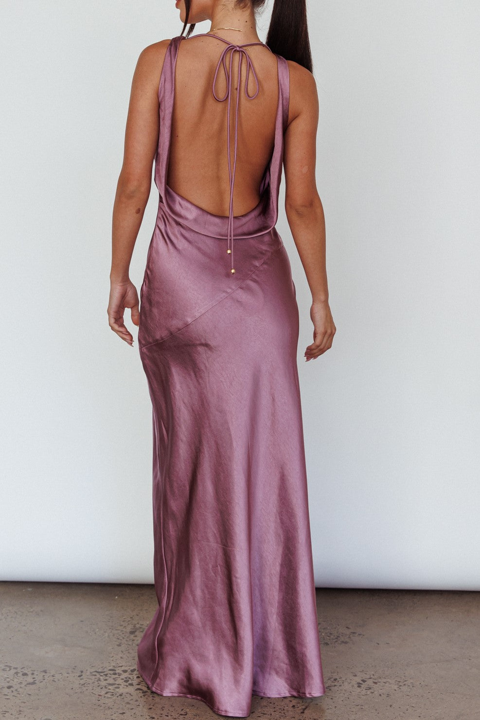 A woman stands with her back facing the viewer, wearing a long, lavender satin dress that features a deep back cut and a tied closure at the neck, in a simple indoor setting.