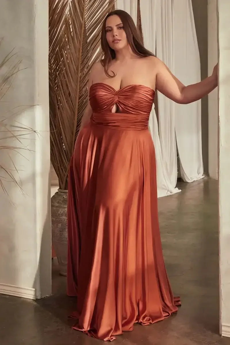 A woman stands elegantly in a flowing, strapless brown dress with a knotted bodice. She poses against a softly lit backdrop featuring draped fabrics and tropical foliage, exuding grace and confidence.
