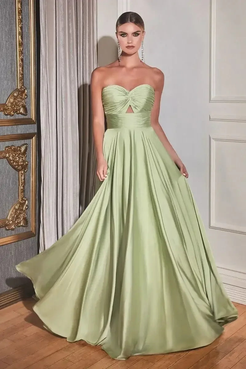 A woman stands elegantly in a flowing, light green gown with a strapless design and a keyhole cutout. The backdrop features ornate decor and soft, draped curtains, enhancing the sophisticated ambiance.
