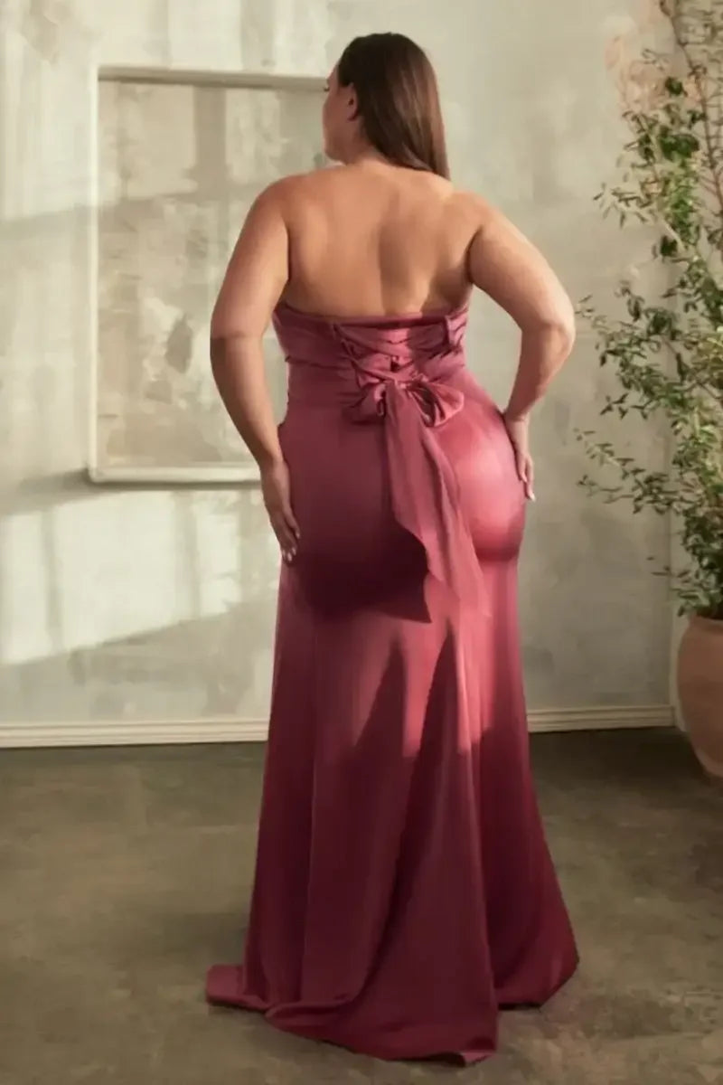 A woman is standing with her back to the viewer, wearing a strapless burgundy dress with a tied bow at the back. She is in a softly lit, minimalist room.