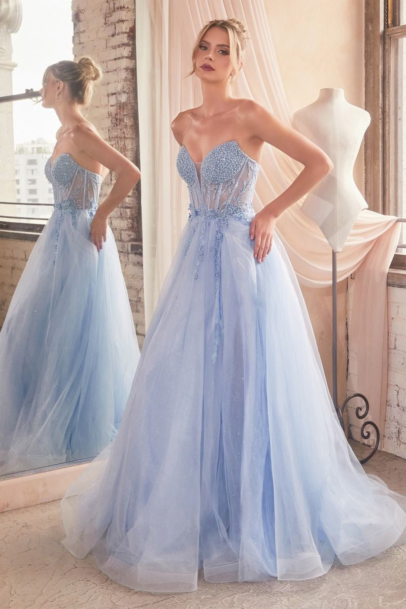 A woman poses in a light blue, strapless gown adorned with beads and tulle, reflecting in a mirror. Sunlight filters through a window, illuminating a vintage-styled room.