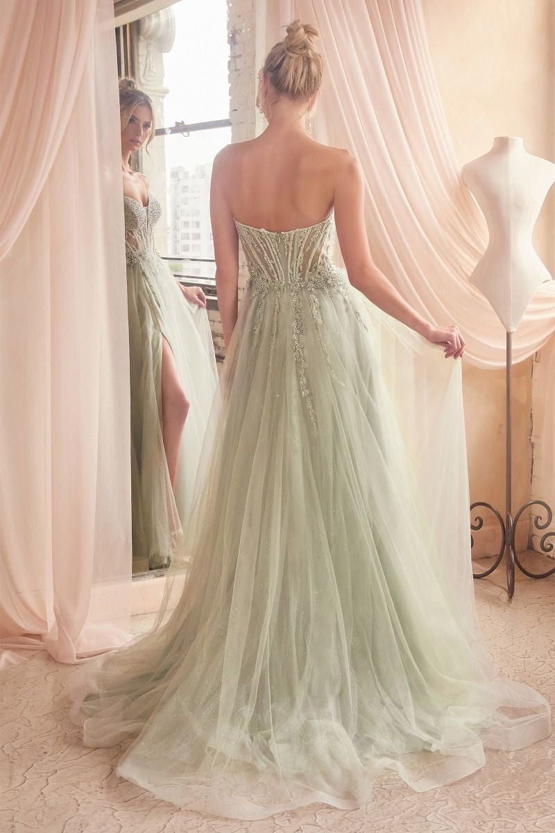 A woman in a flowing, pale green gown stands before a mirror, gently holding the fabric as she gazes at her reflection in a softly lit room with sheer curtains and a dress form nearby.