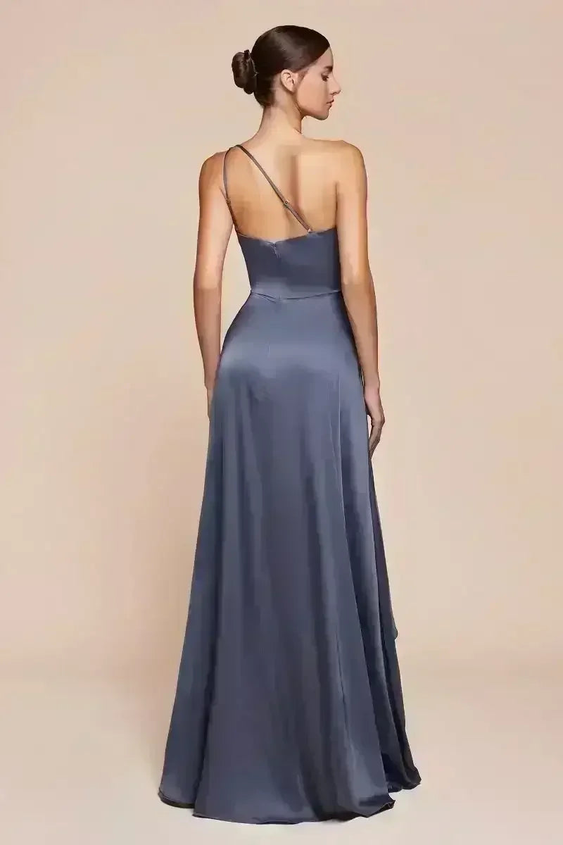 A woman stands gracefully with her back turned, wearing a long, flowing gray satin dress. The backdrop is a smooth, neutral-colored surface, emphasizing her elegant silhouette.