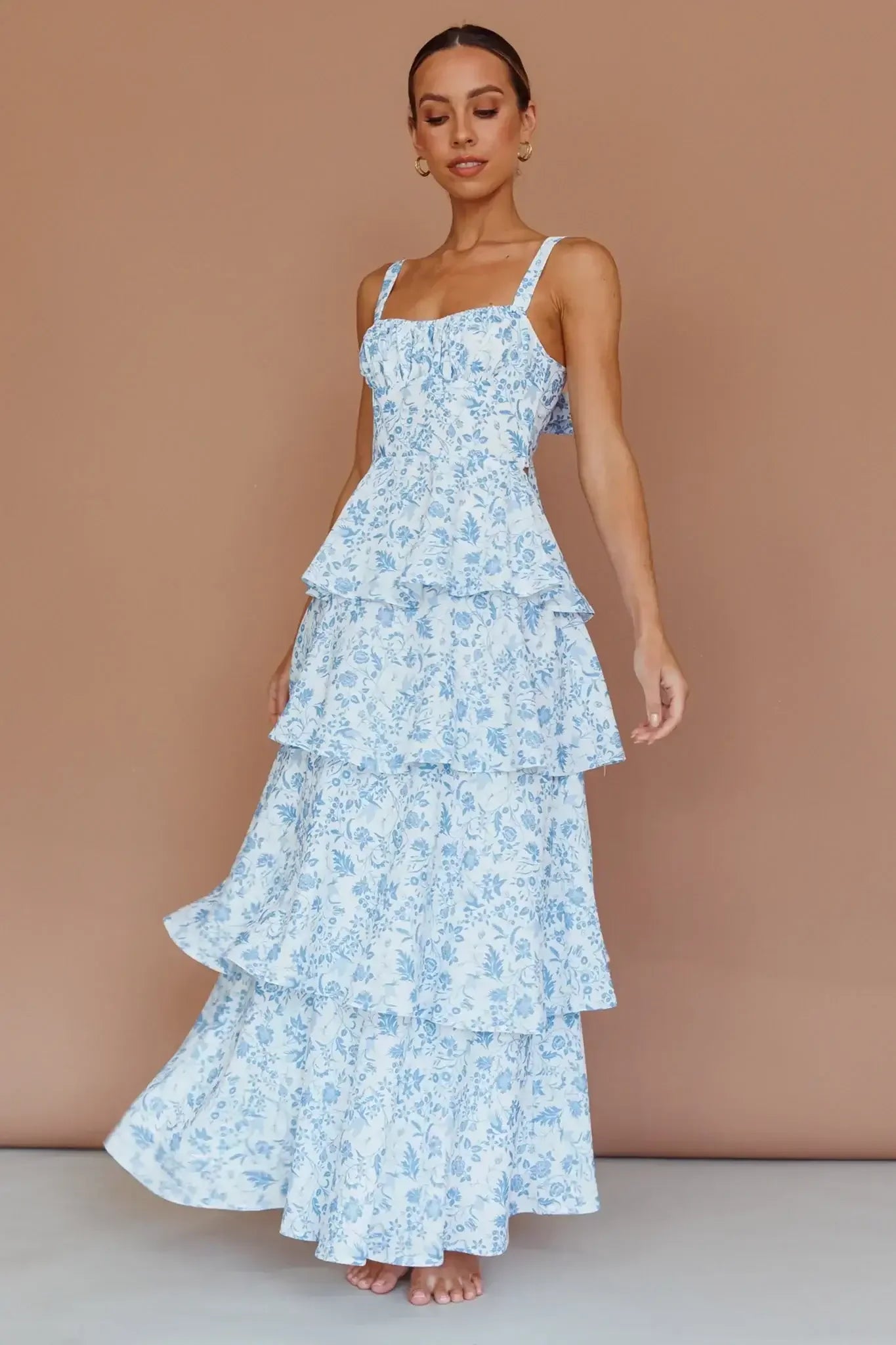A woman stands gracefully in a blue and white floral dress with layered ruffles. She appears to be posing against a warm, neutral background, showcasing the dress's elegant design.
