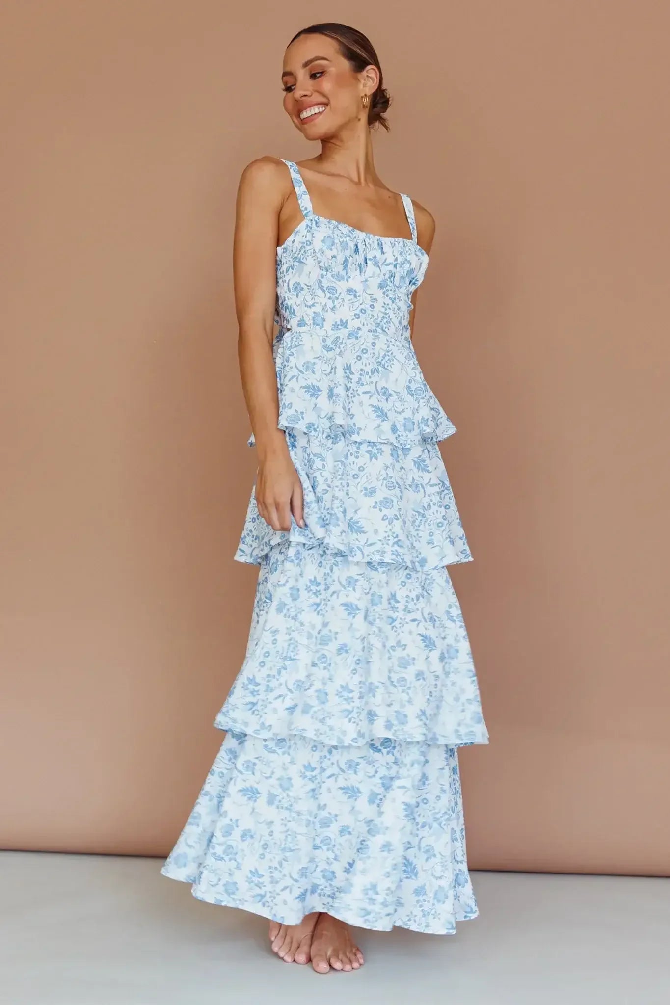 A woman is smiling while wearing a blue floral tiered dress. She stands barefoot on a textured surface against a plain brown backdrop, conveying a warm and cheerful vibe.