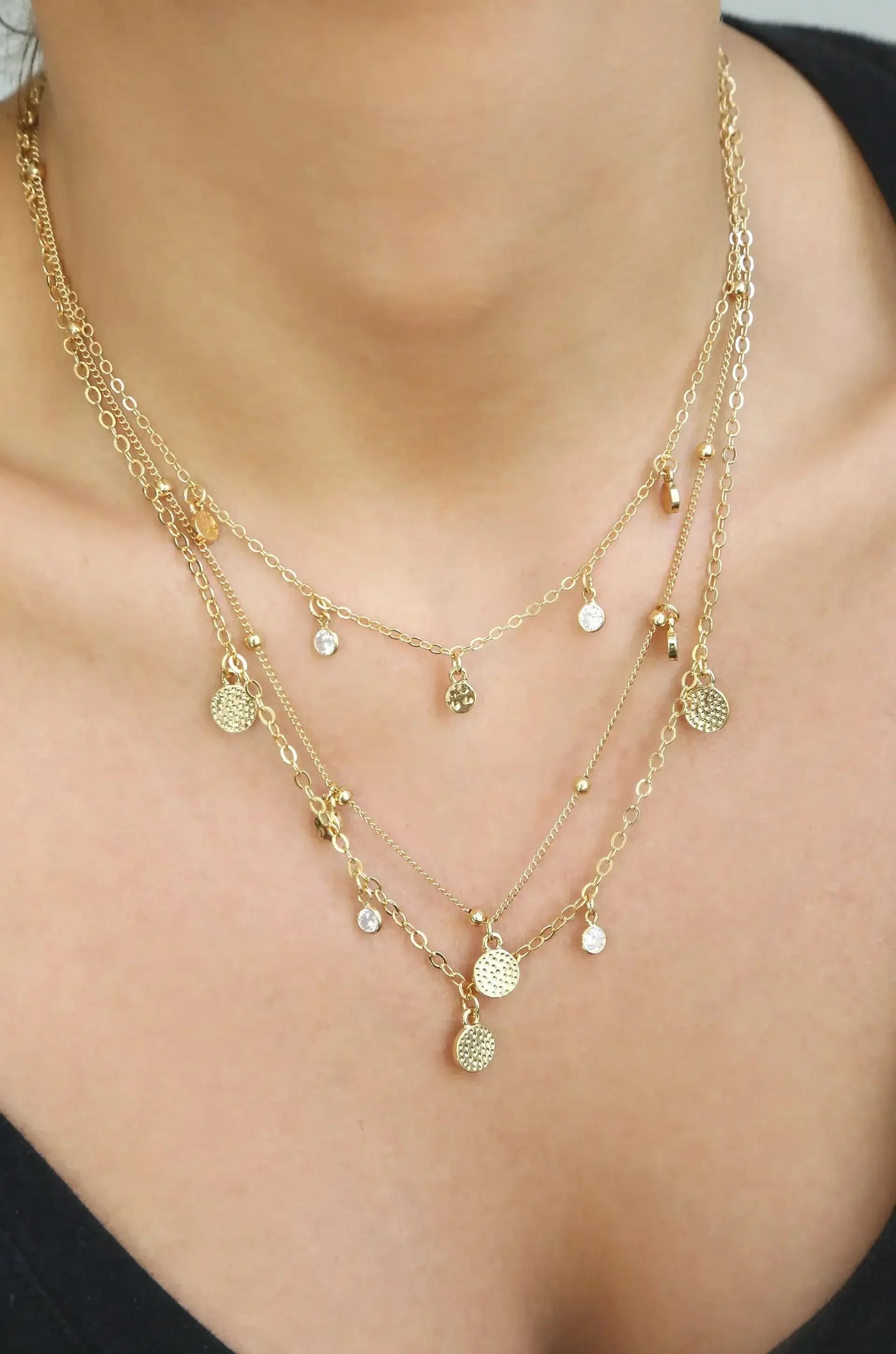 A layered gold necklace adorned with small charms and clear crystals rests on a person’s neck, enhancing the collarbone above a dark top in a softly lit setting.