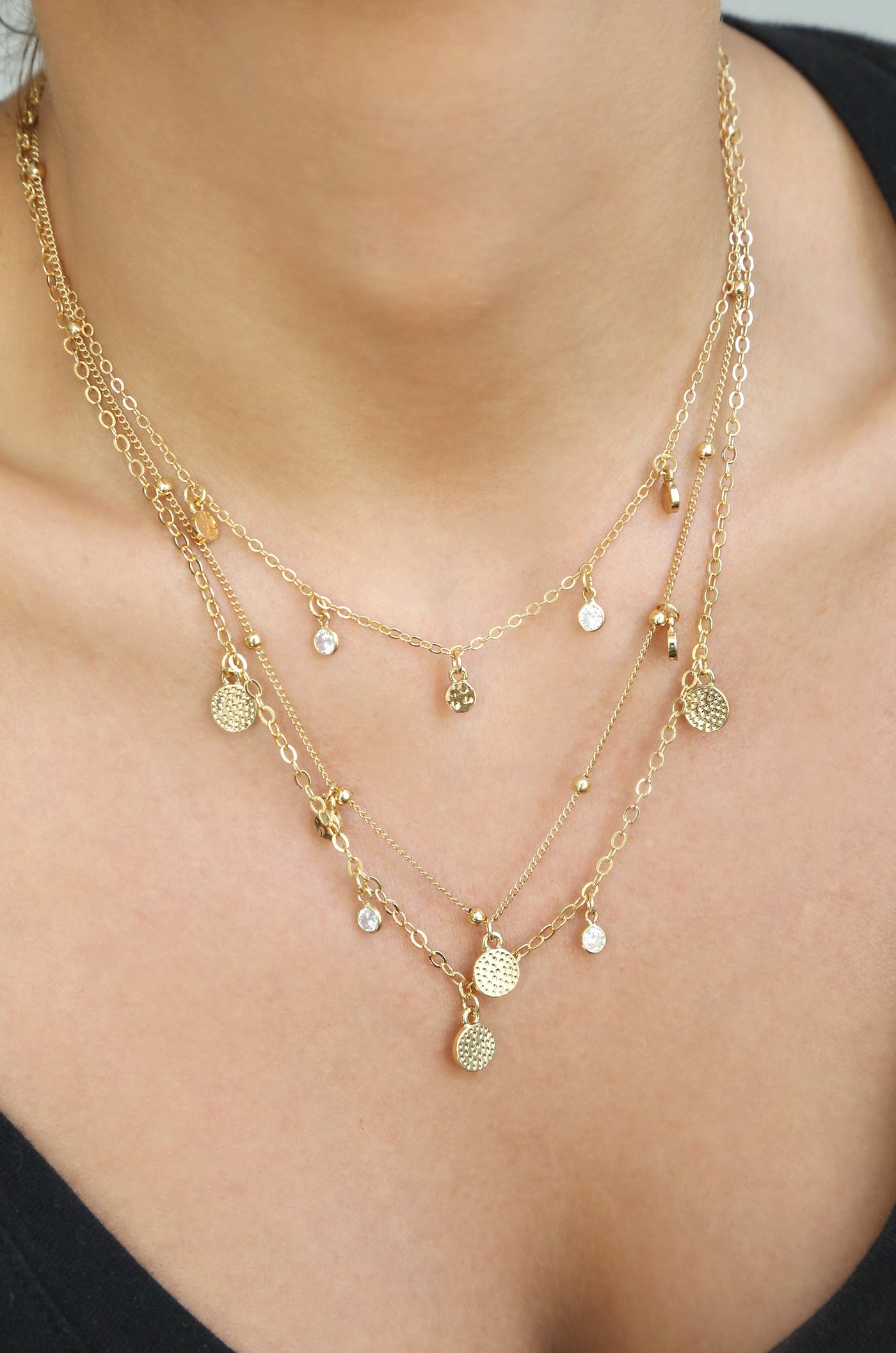 A layered gold necklace adorned with small charms and clear crystals rests on a person’s neck, enhancing the collarbone above a dark top in a softly lit setting.