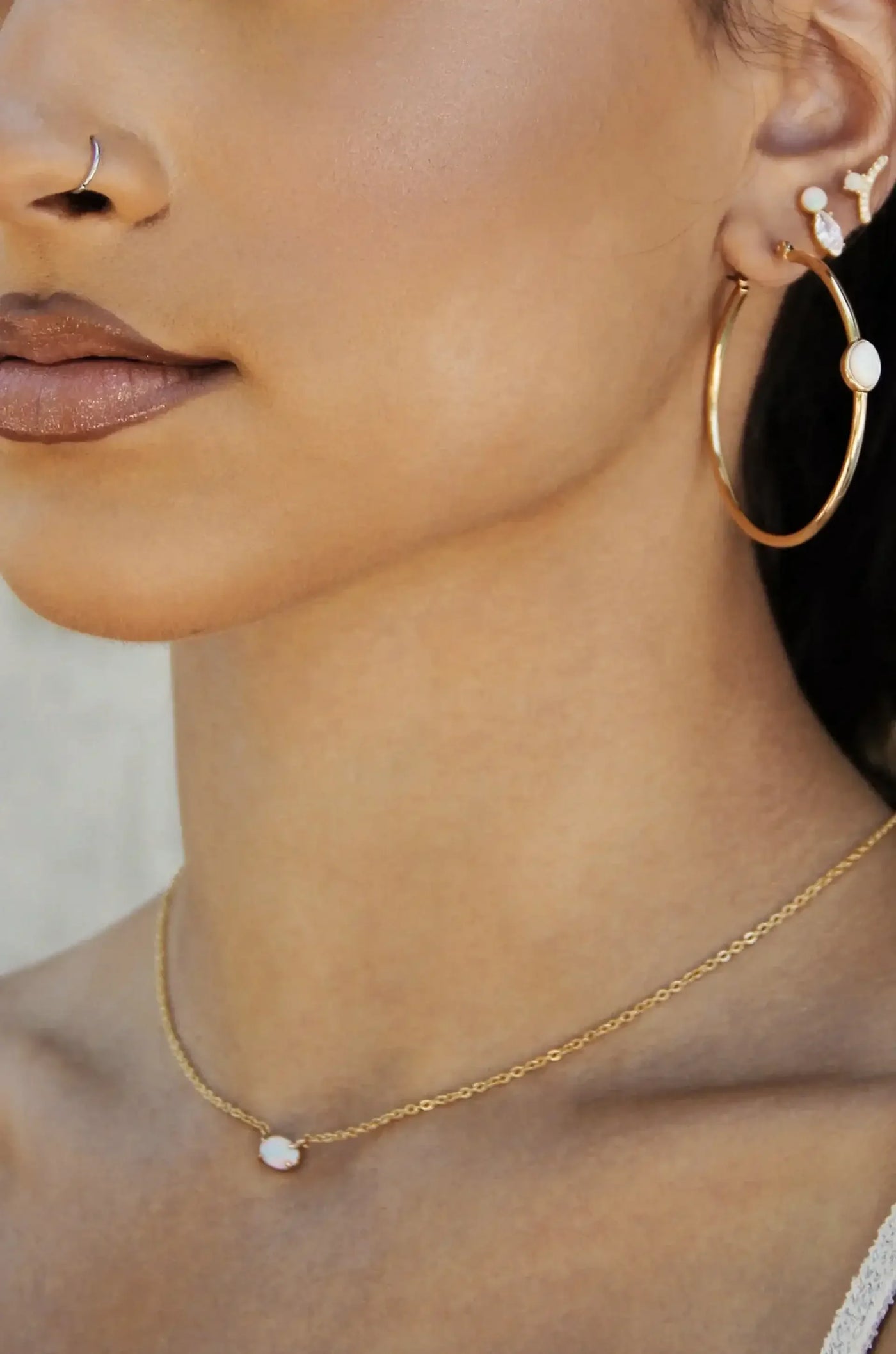 A close-up of a woman’s profile showcases her gold hoop earrings and a delicate gold necklace with an opal pendant, set against a softly blurred background.