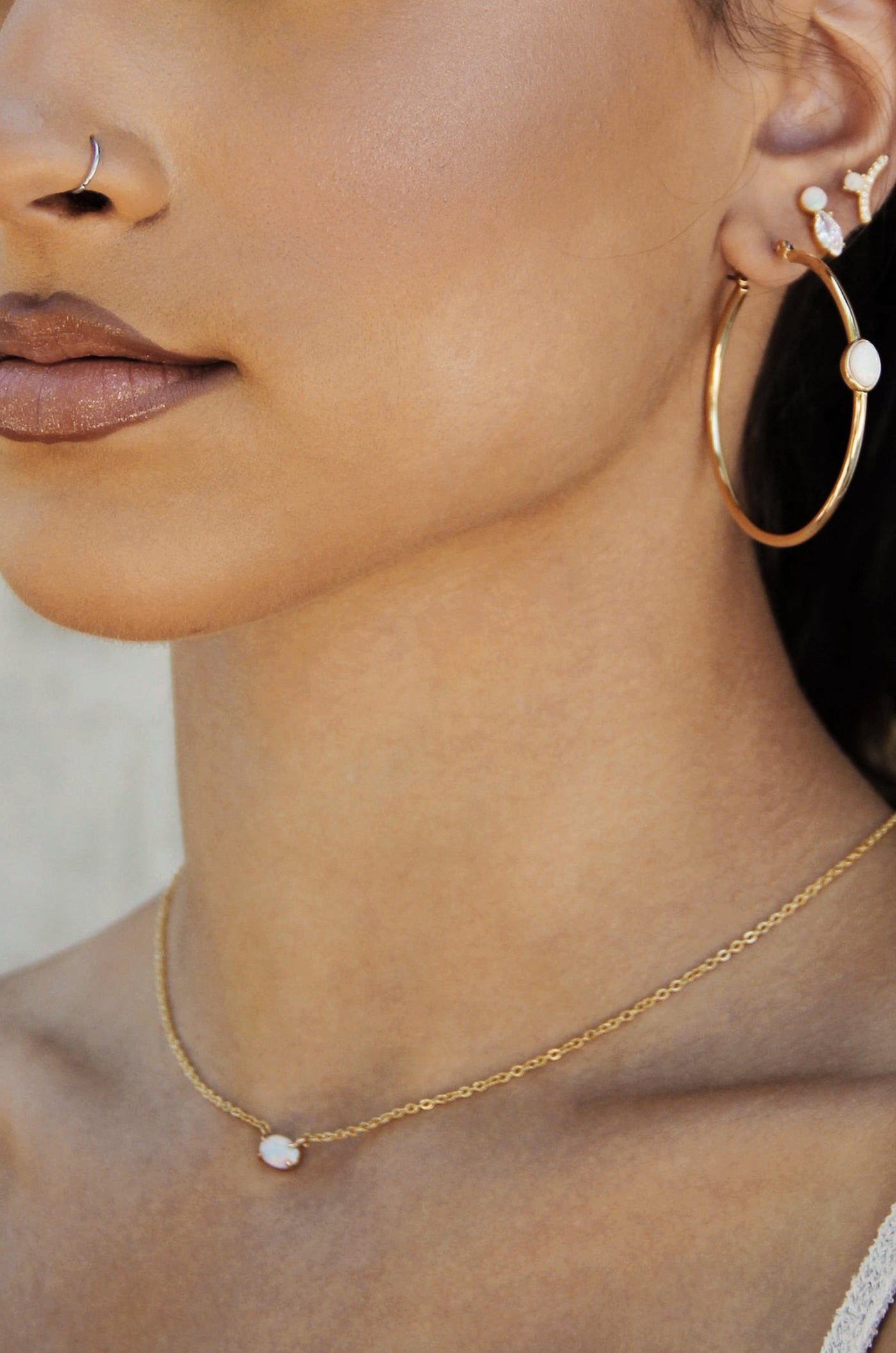 A close-up of a woman’s profile showcases her gold hoop earrings and a delicate gold necklace with an opal pendant, set against a softly blurred background.