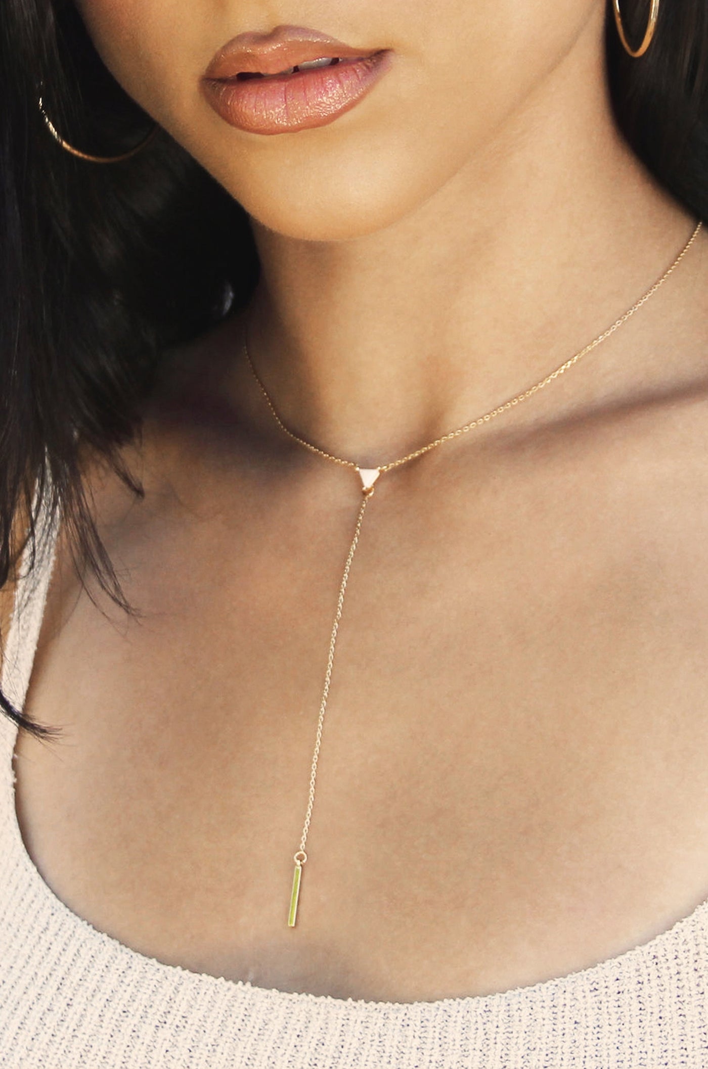 A delicate gold necklace adorns a woman's neck, featuring a small triangular pendant at the center and a thin chain dropping down with a slender, vertical green accent. The background is softly blurred.