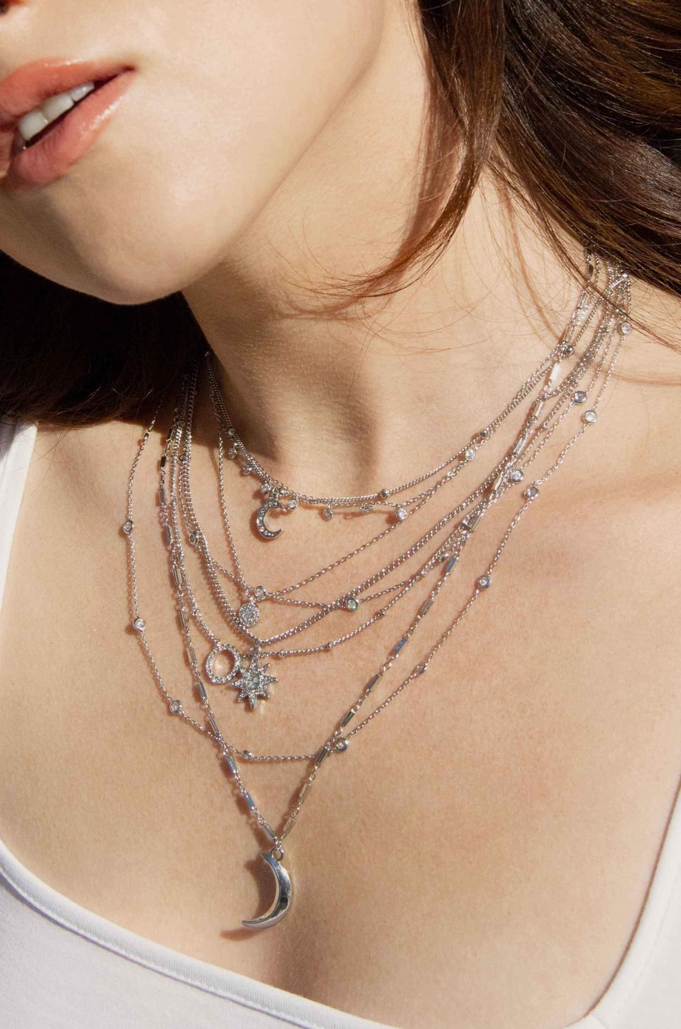 Layered silver necklaces with various decorative charms hang gracefully around a person's neck, catching the light against soft skin, set in a bright, natural light environment.