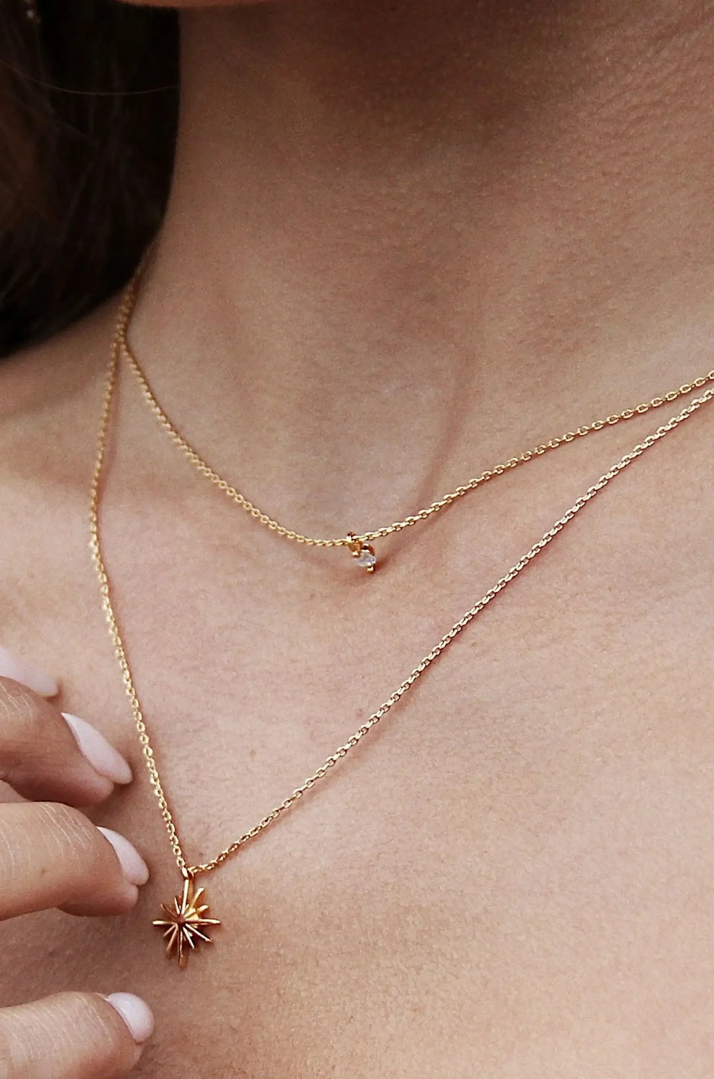 A gold chain necklace with a star-shaped pendant rests on a neck. A hand gently touches the pendant, emphasizing its delicate design against smooth skin.