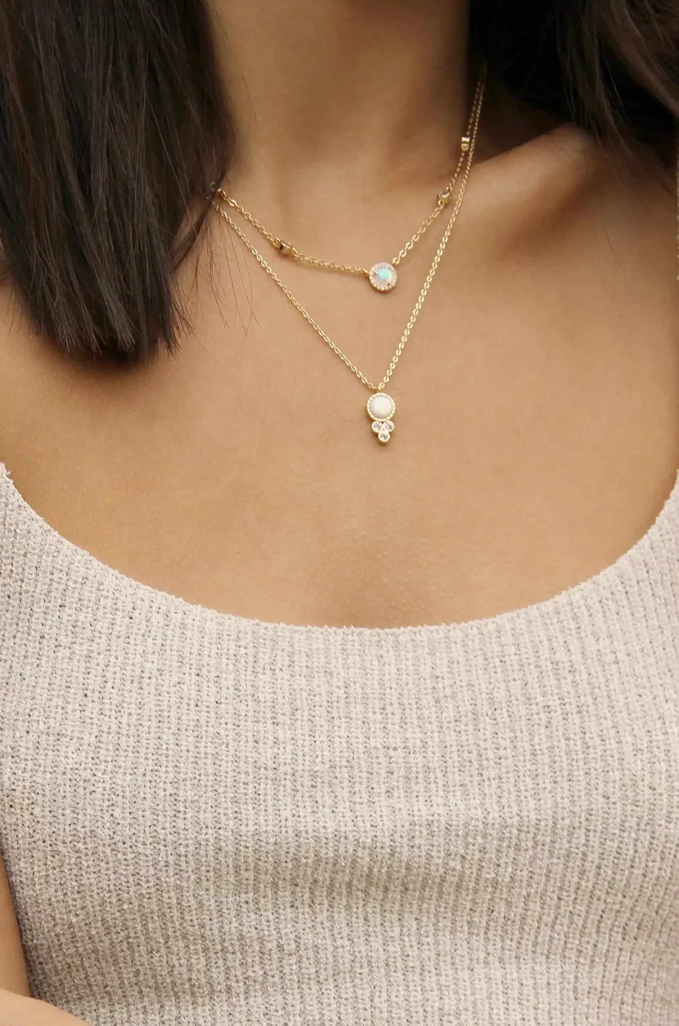 Delicate gold necklaces layer around a person's neckline. One features a luminous opal, while the other showcases a round pearl pendant, set against a textured beige top.