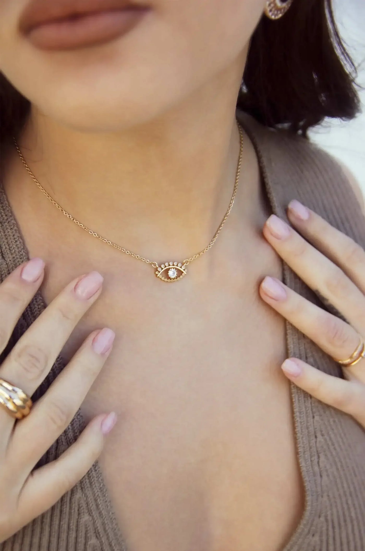 A gold necklace featuring an eye pendant sits against a person's neck, while their fingers gently touch the necklace. The background is softly blurred, emphasizing the jewelry.