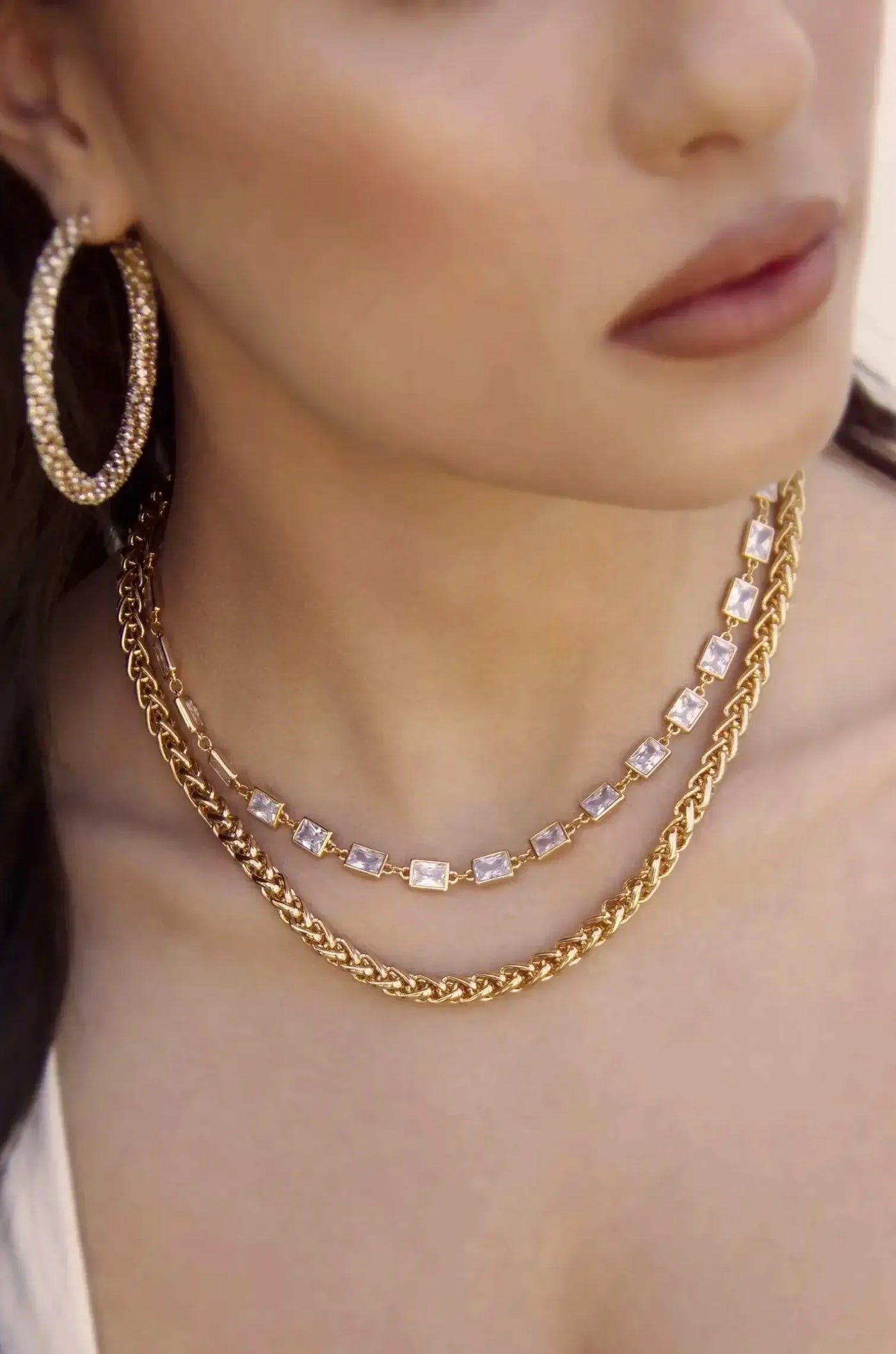A person wears layered gold necklaces with sparkling pendants and large hoop earrings. The focus is on the jewelry, accentuating the neckline against a soft, neutral background.