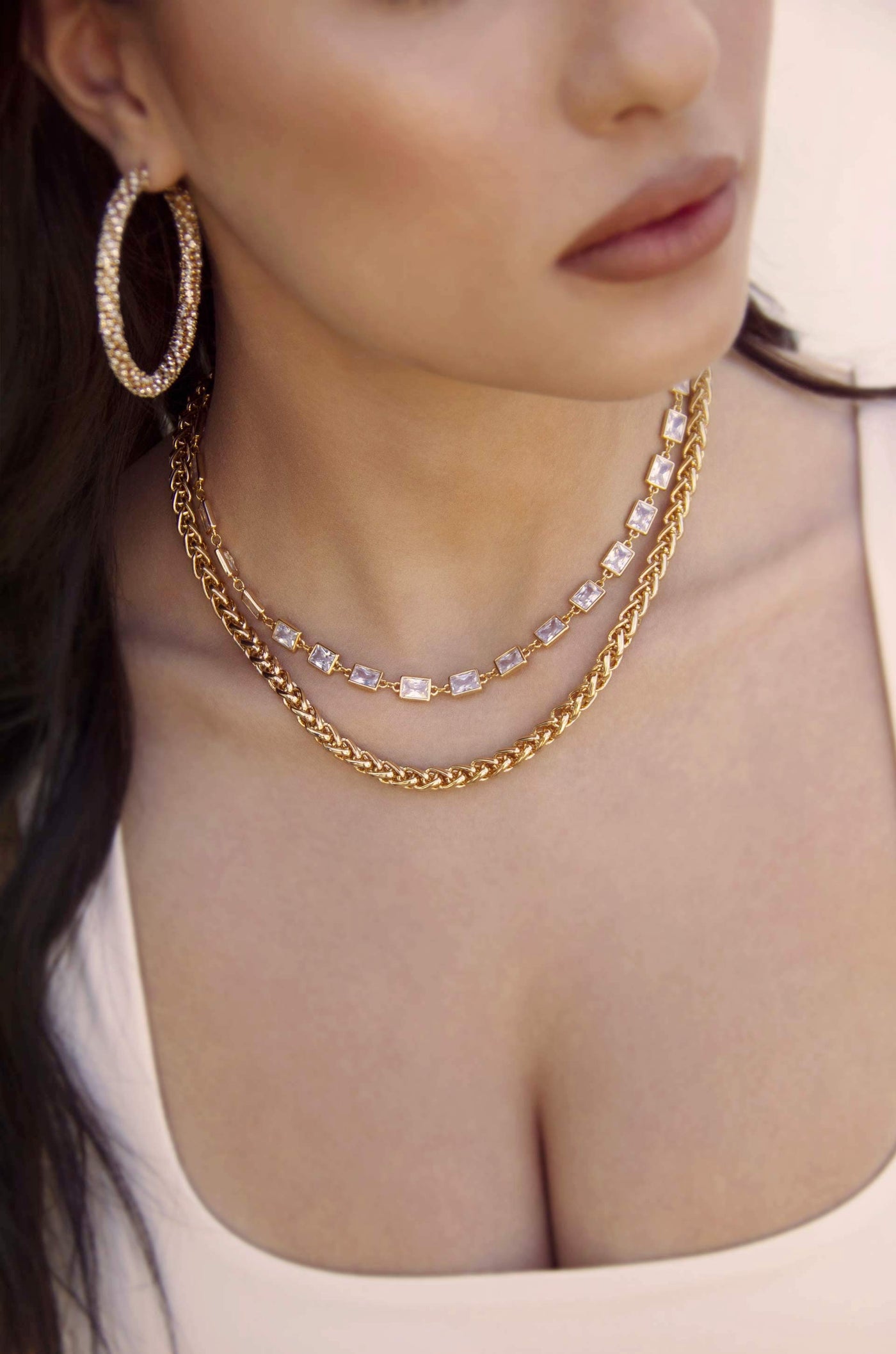 A person wears layered gold necklaces with sparkling pendants and large hoop earrings. The focus is on the jewelry, accentuating the neckline against a soft, neutral background.