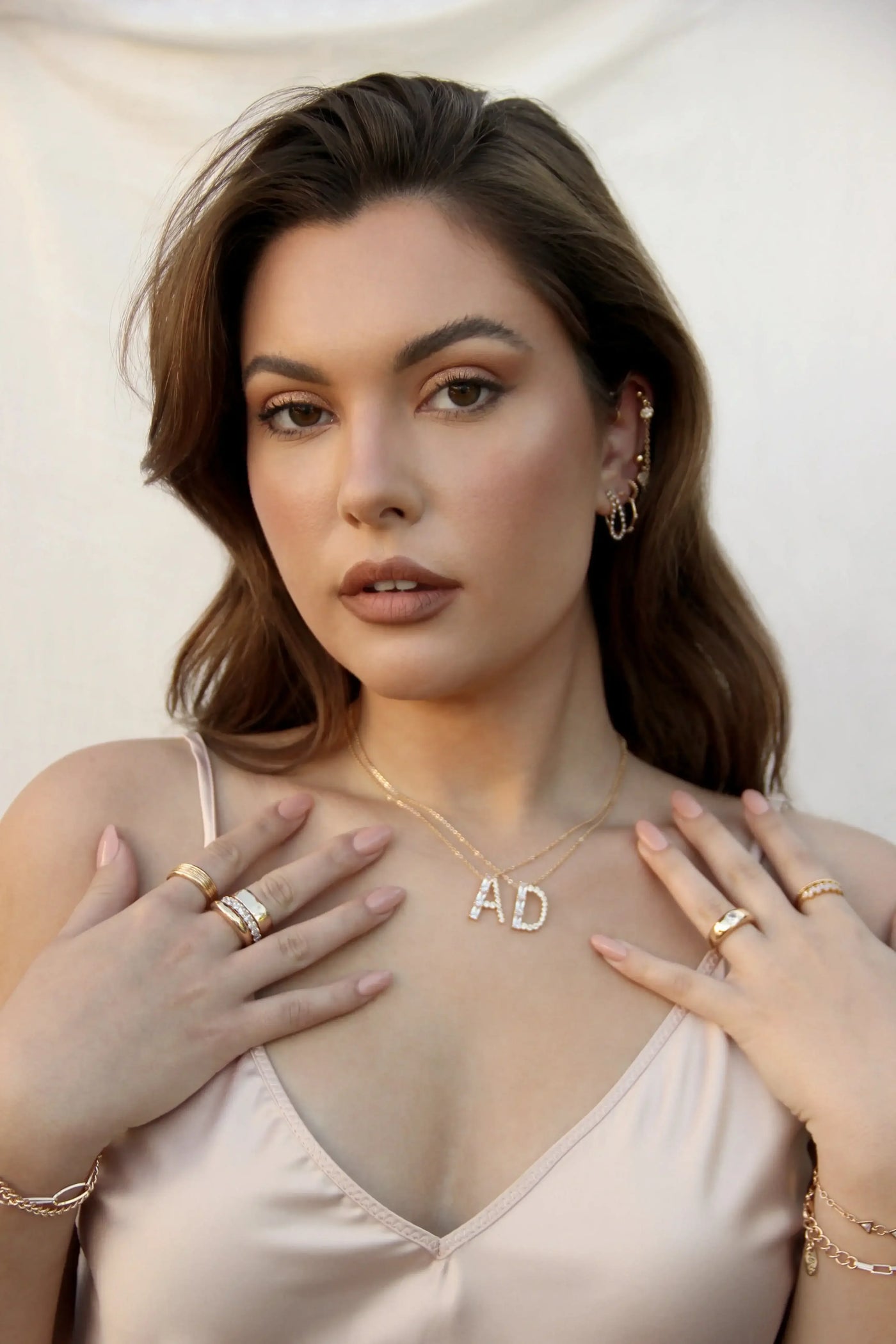 A woman with long, wavy hair wears a satin top and showcases jewelry, including a sparkling "AD" necklace, while gently touching her shoulders against a soft, neutral background.