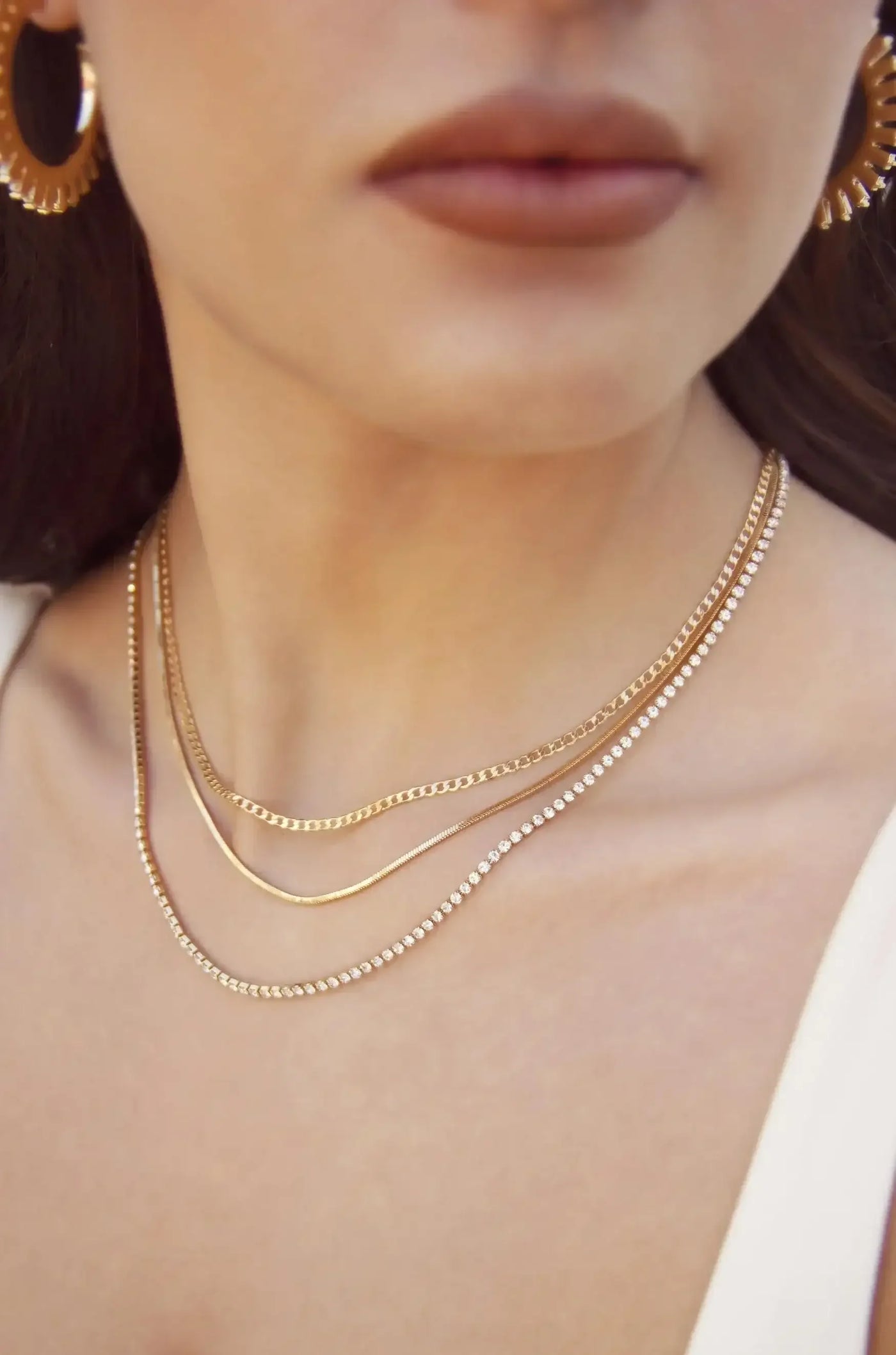 Layered necklaces adorn a woman's neck, gleaming in the light. She has a subtle expression, with golden hoop earrings visible. The background is softly blurred, emphasizing the jewelry.