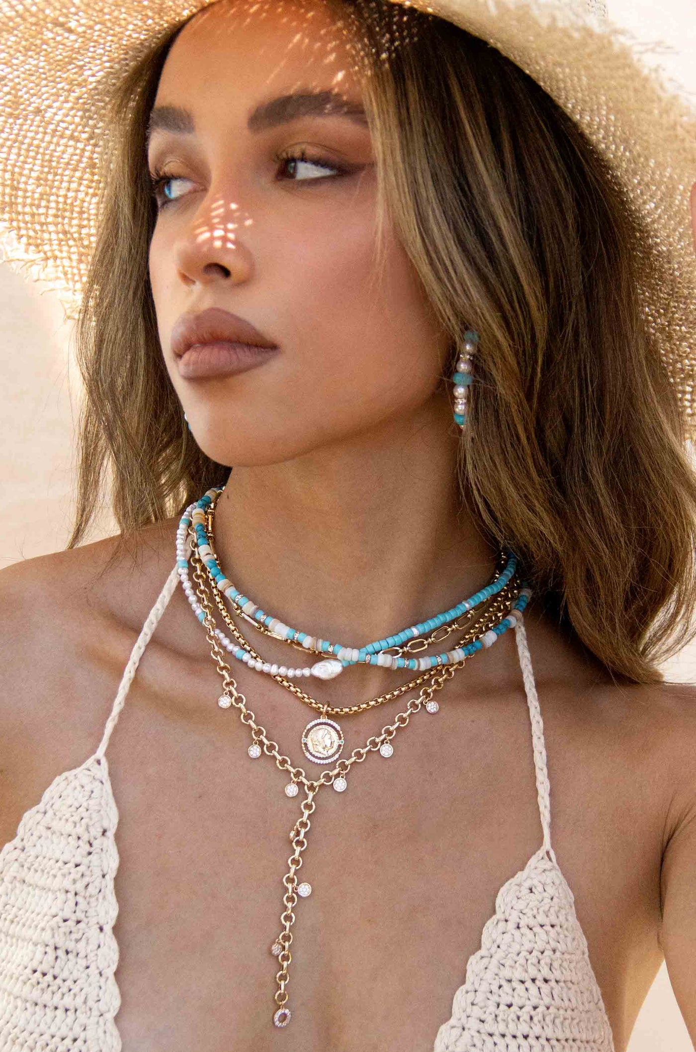 A woman wears layered necklaces featuring turquoise beads, pearls, and gold chains, set against a neutral background. Sunlight creates intricate patterns on her face and neck, enhancing the warm scene.