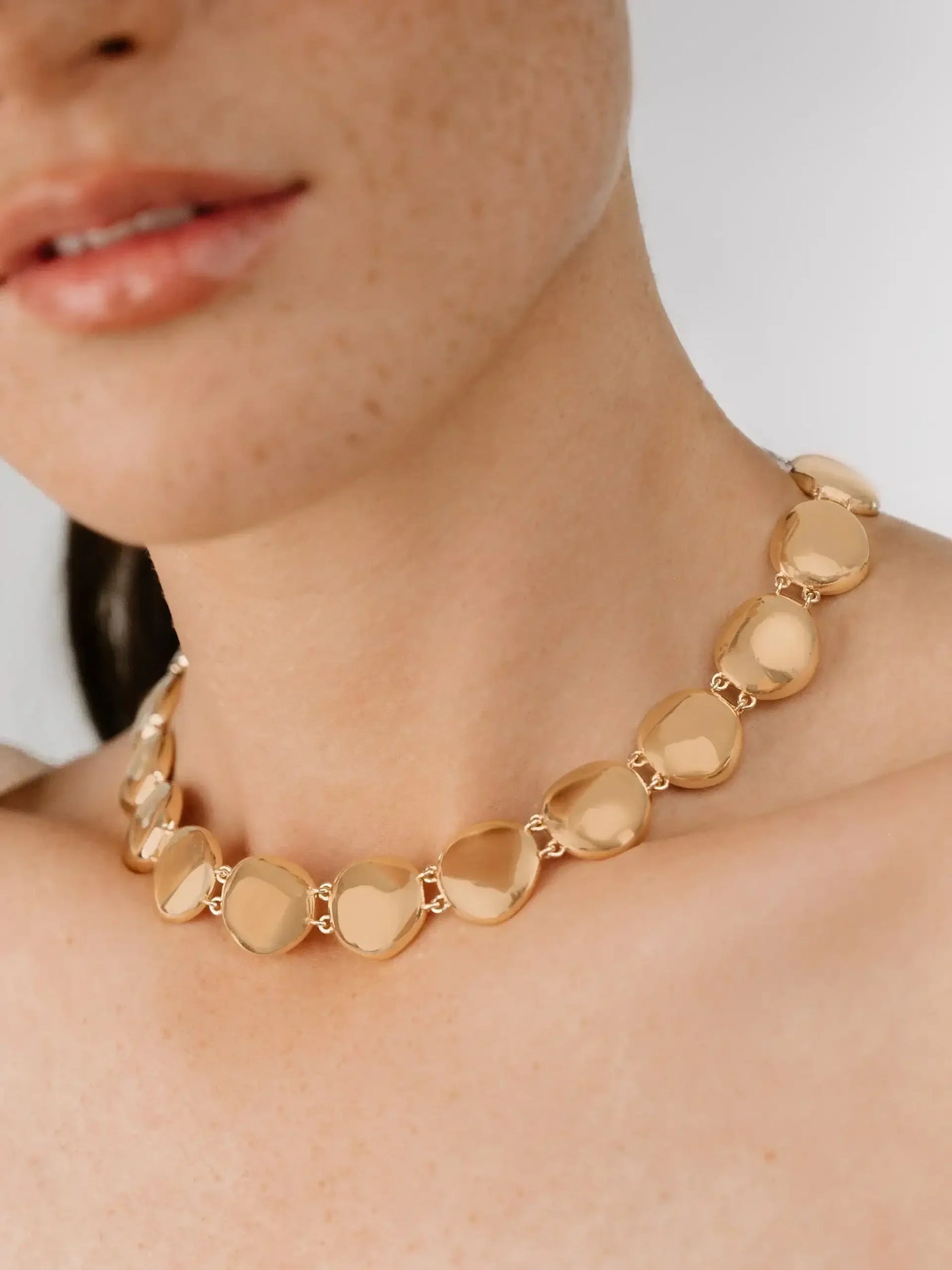 A golden heart-shaped necklace rests elegantly around the neck of a person with light freckles, complemented by smooth skin and a neutral background. The expression is serene and natural.