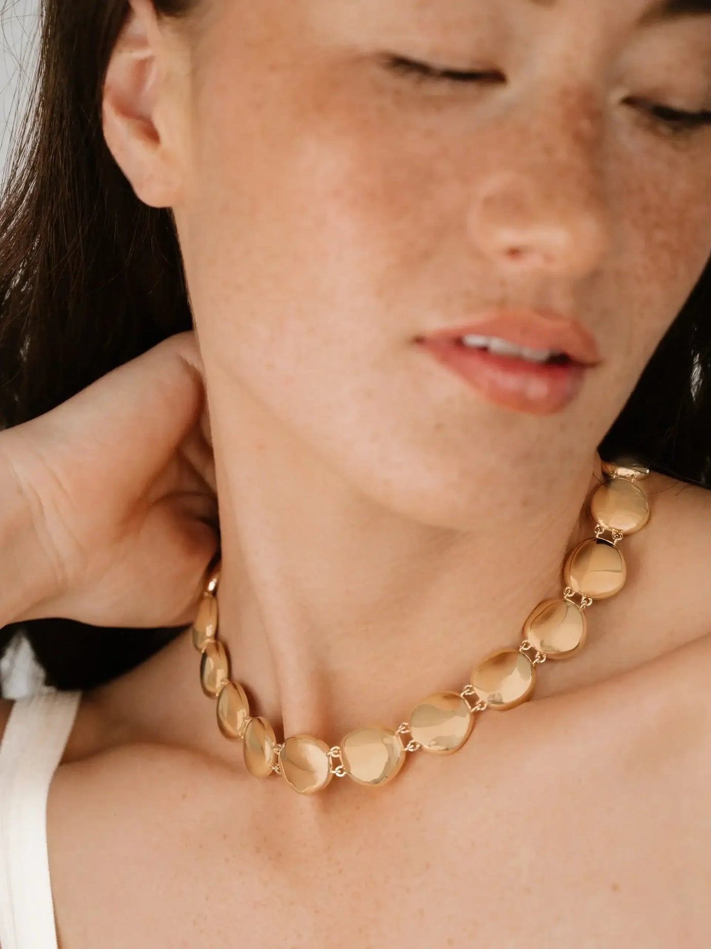A woman is gently adjusting a gold necklace adorned with round, shiny links. She has a relaxed expression, her head slightly turned against a softly blurred background, highlighting her features.
