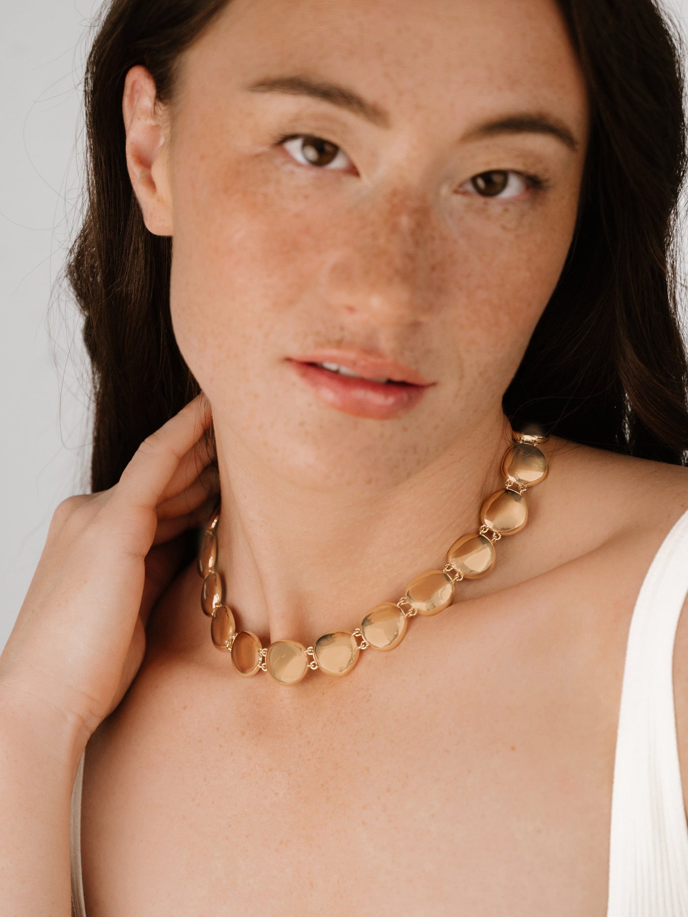 A woman with light skin and freckles gently touches a gold necklace around her neck, showcasing the smooth, rounded links. She has long, dark hair and a subtle expression.