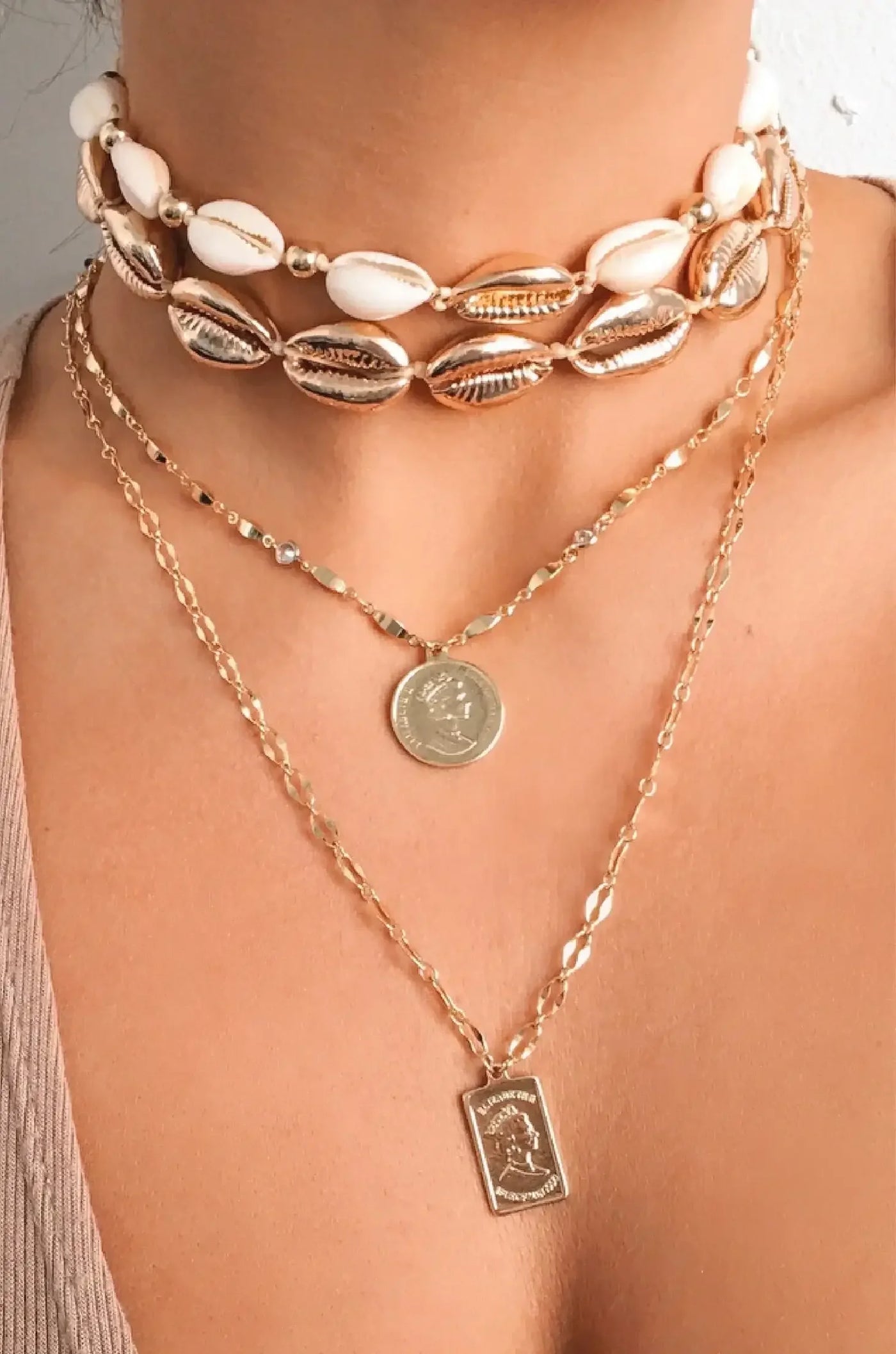 A layered necklace design featuring a choker made of shells and gold, alongside two gold chains; one adorned with a coin and the other with a rectangular pendant, worn on bare skin.
