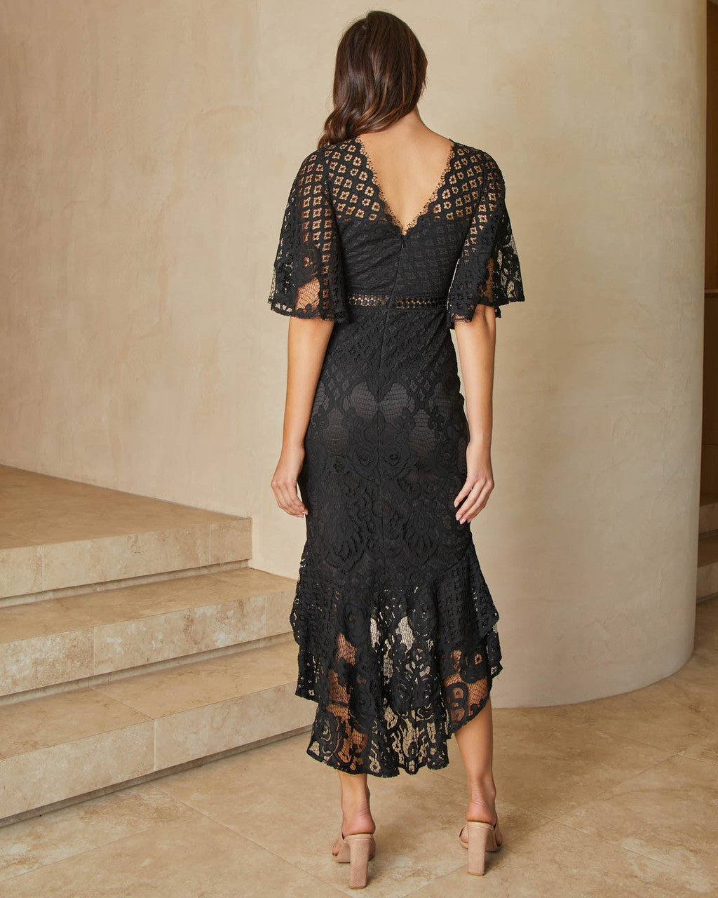 A woman wears a black lace dress with bell sleeves, standing on stairs in a minimalistic, light-colored interior, showcasing the intricate patterns of the fabric and a stylish, flowy hem.