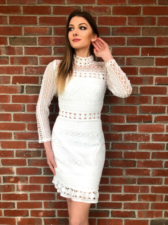 A woman stands elegantly against a brick wall, wearing a stylish white lace dress with a high collar and intricate designs, holding her hair back while gazing thoughtfully.