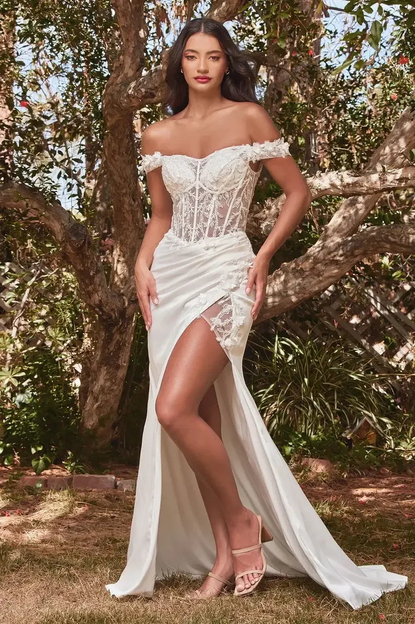 A woman stands elegantly in a white off-the-shoulder dress with lace details, exposing her leg. She is surrounded by greenery and trees, suggesting a natural outdoor setting.